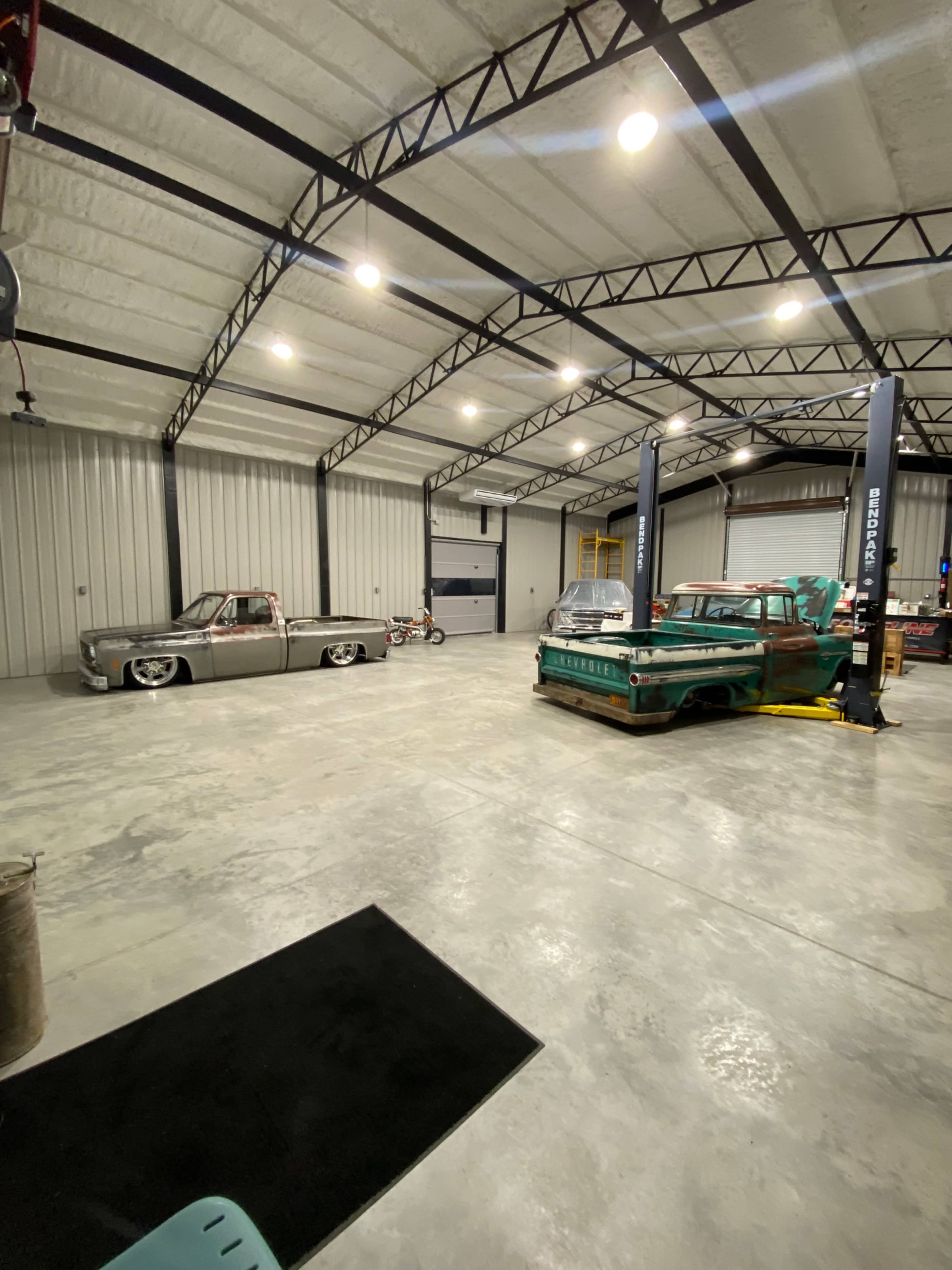 An industrial garage features several classic cars, including a silver pickup and a green vehicle, with a vehicle lift in the center and concrete flooring.