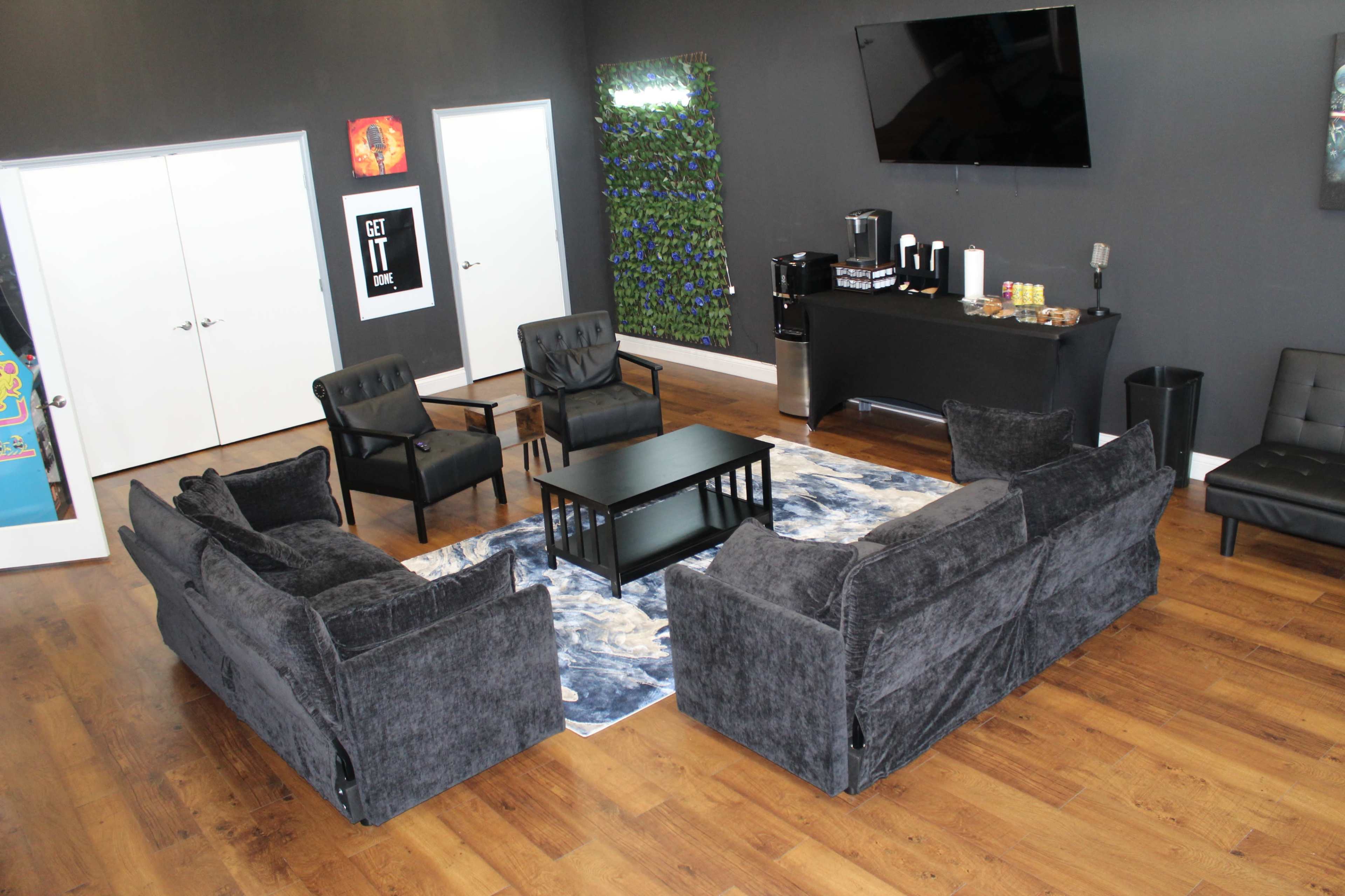 The image shows a modern lounge area featuring four chairs and two sofas arranged around a coffee table on a patterned rug, with a TV mounted on the wall and a refreshment station nearby.