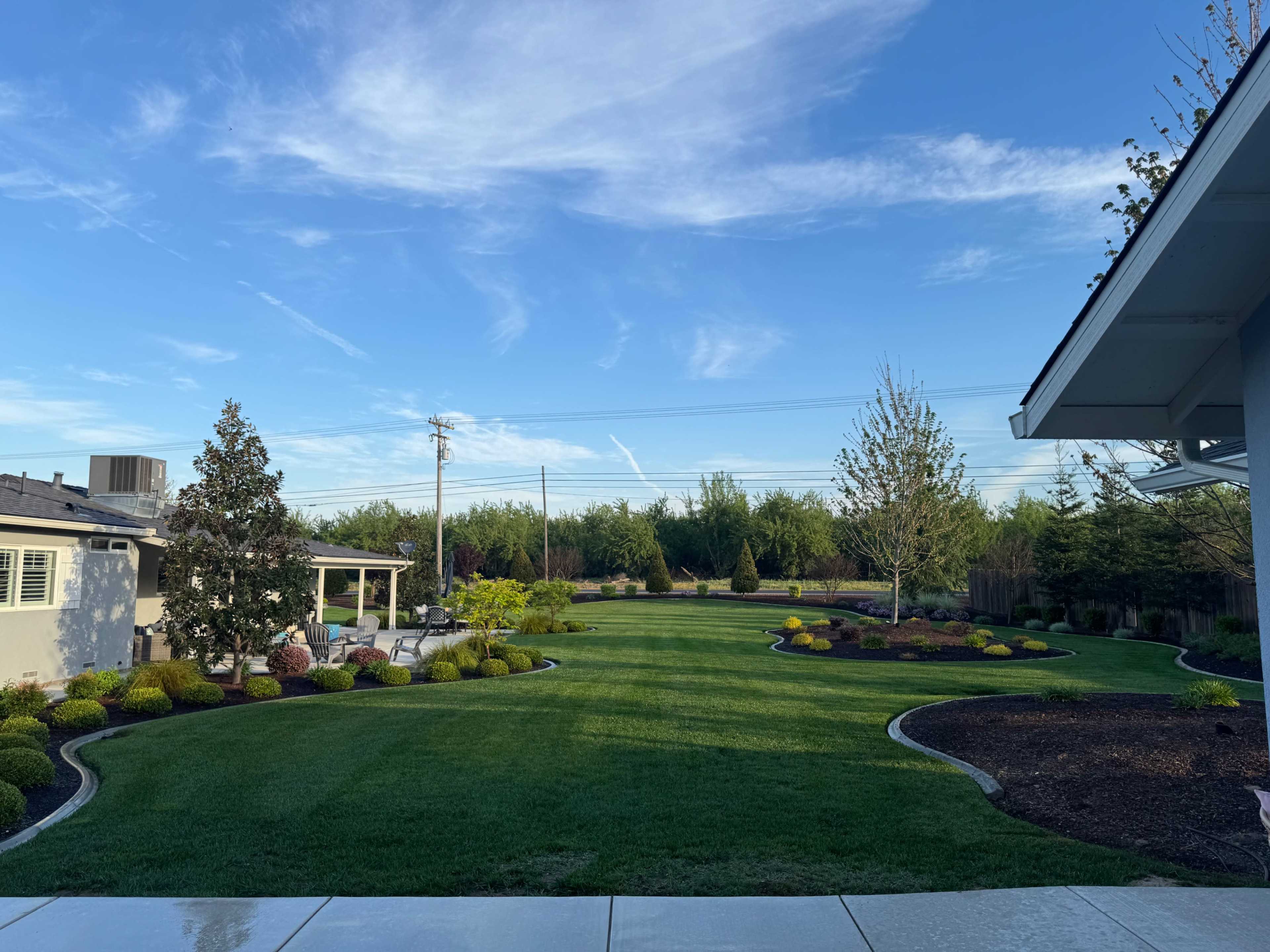 The image shows a well-maintained lawn with neatly trimmed grass, bordered by flower beds and trees under a clear blue sky.