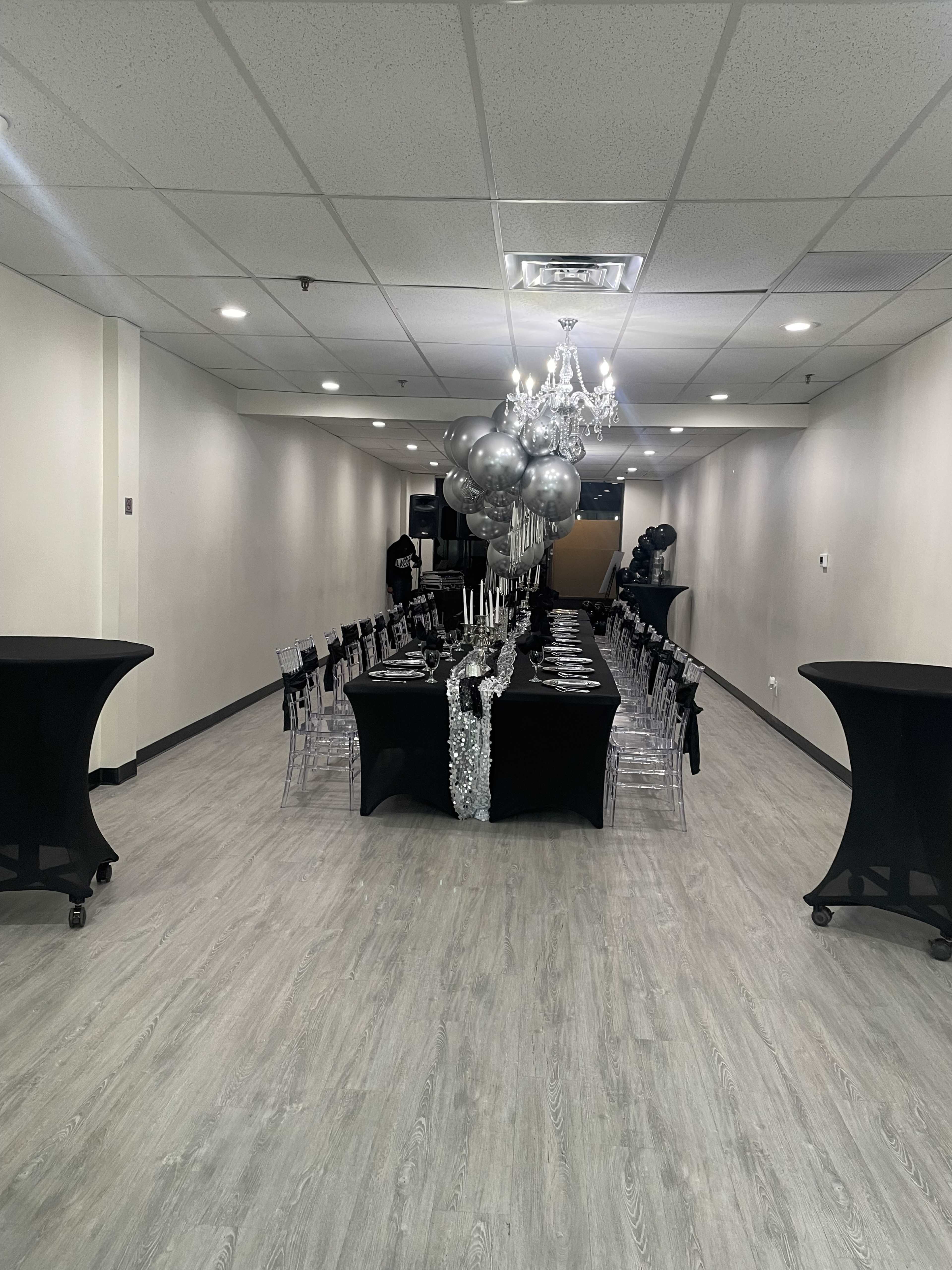 A long dining table is set with black tablecloths and silver decorations in a spacious, well-lit event room.