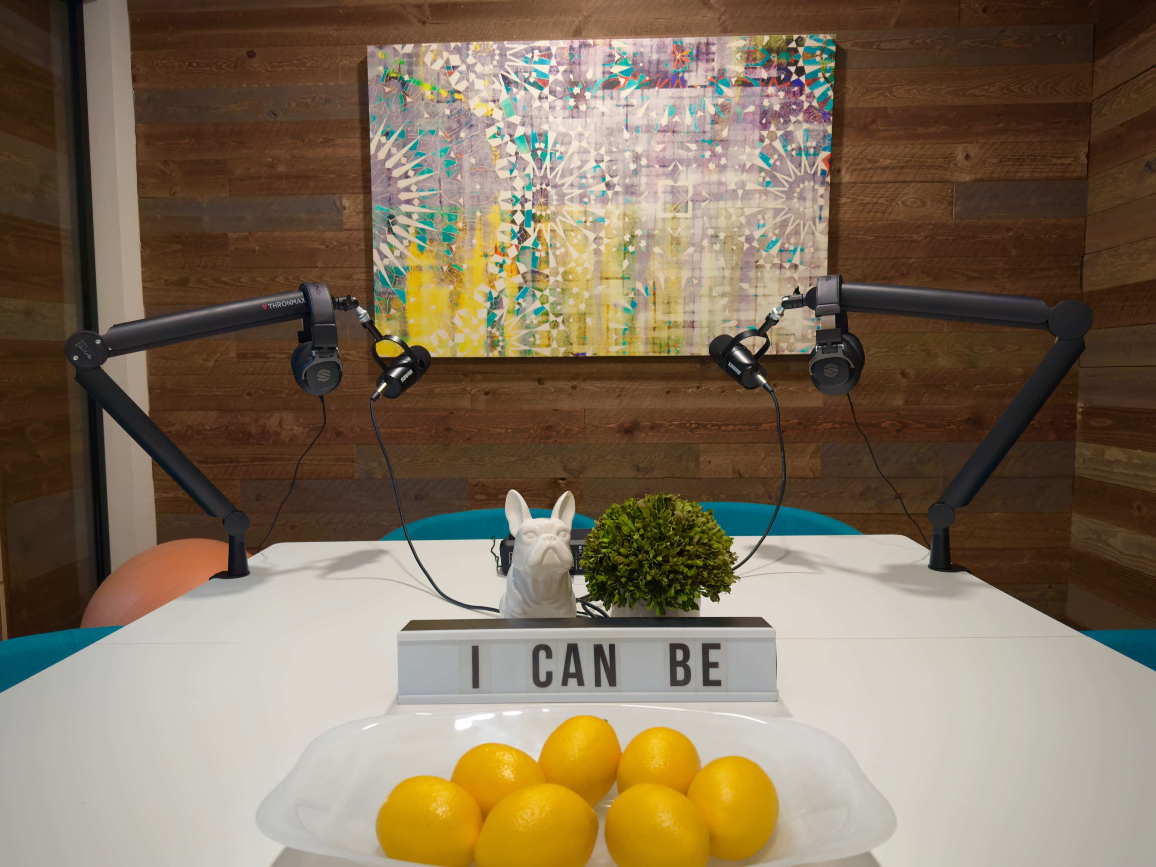 A brightly lit room features a table with a bowl of lemons, a decorative plant, and a sign reading "I CAN BE," against a colorful abstract painting and two microphone stands.