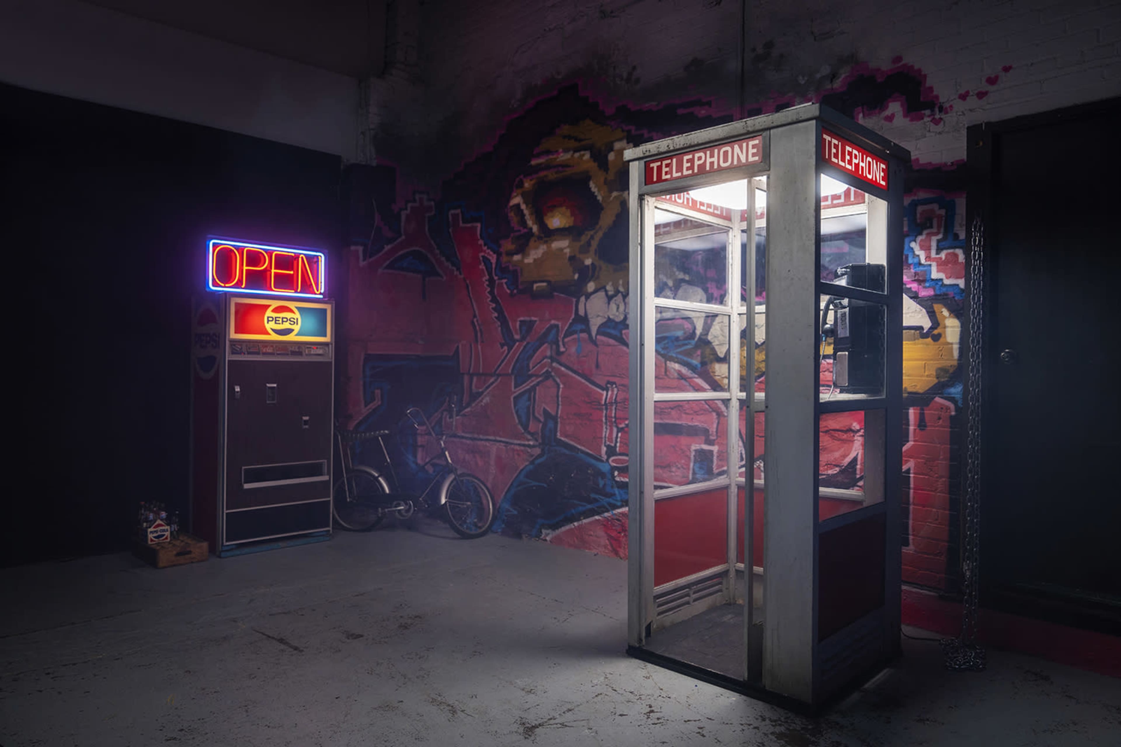 The image shows a retro telephone booth and a vintage Pepsi vending machine set against a graffiti-covered wall in a dark room.