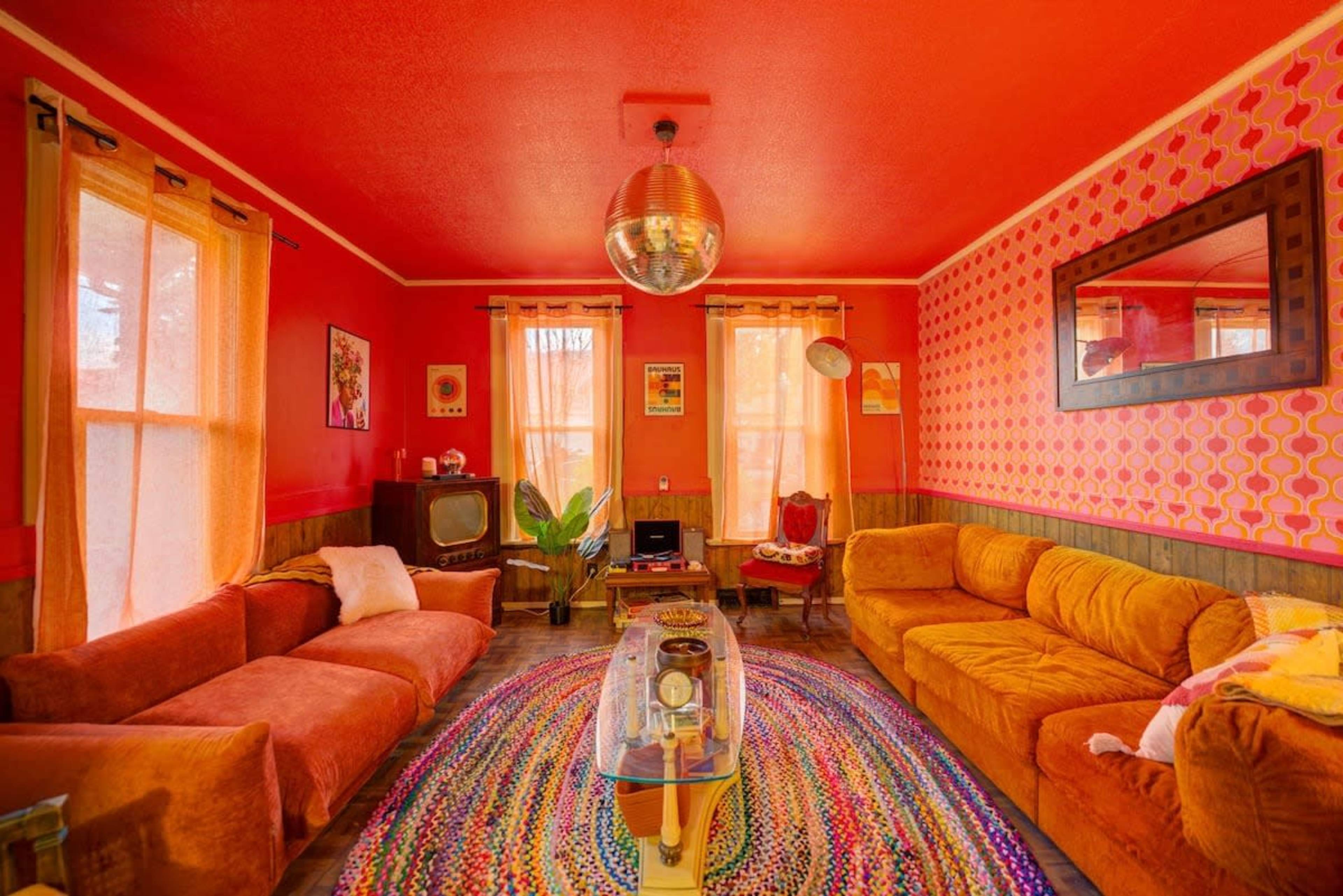 The room features a vibrant red interior with vintage furniture, a disco ball, and a colorful rug at the center.