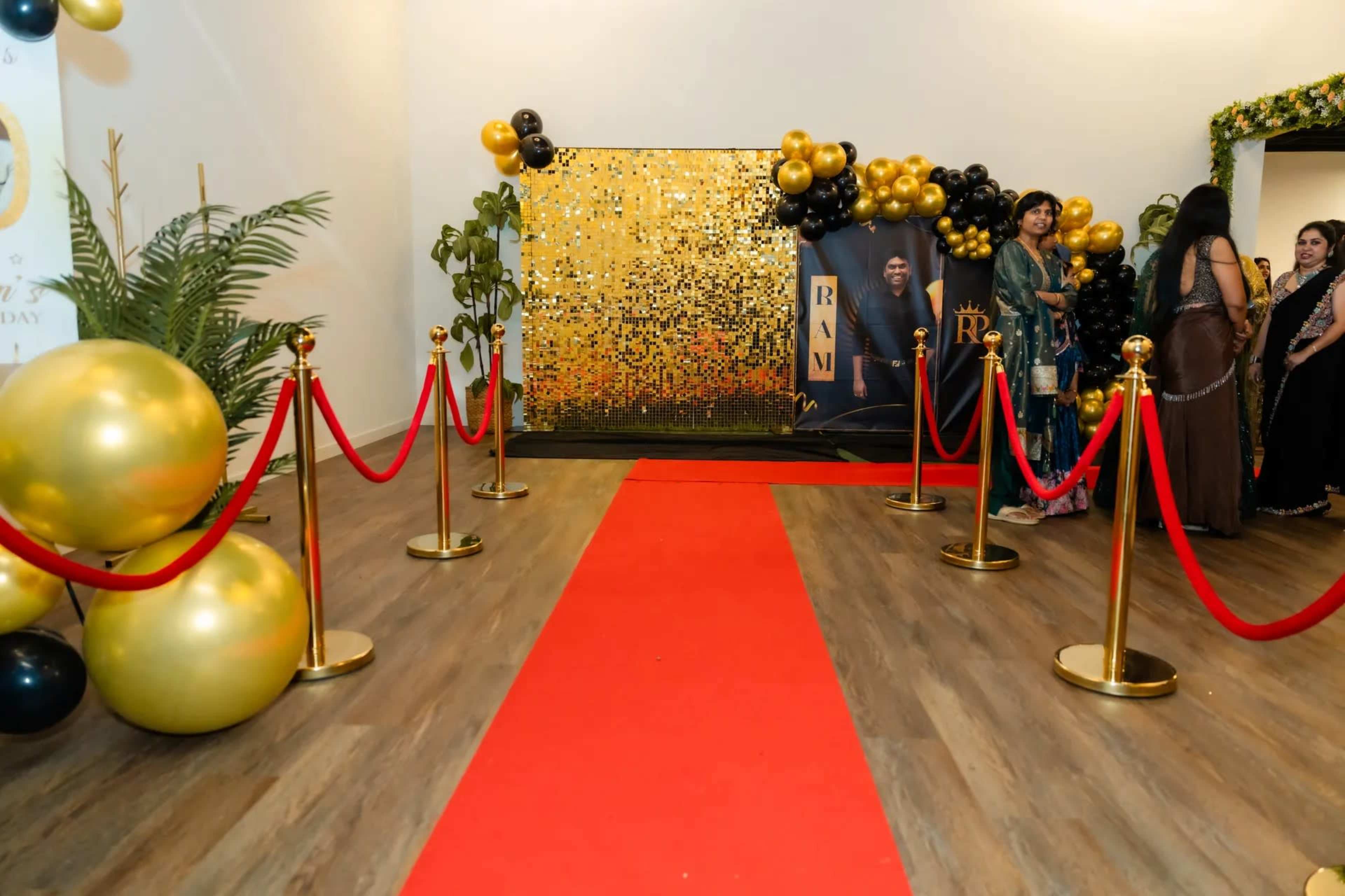A red carpet leads to a sparkling backdrop decorated with gold and black balloons, flanked by velvet ropes and potted plants.