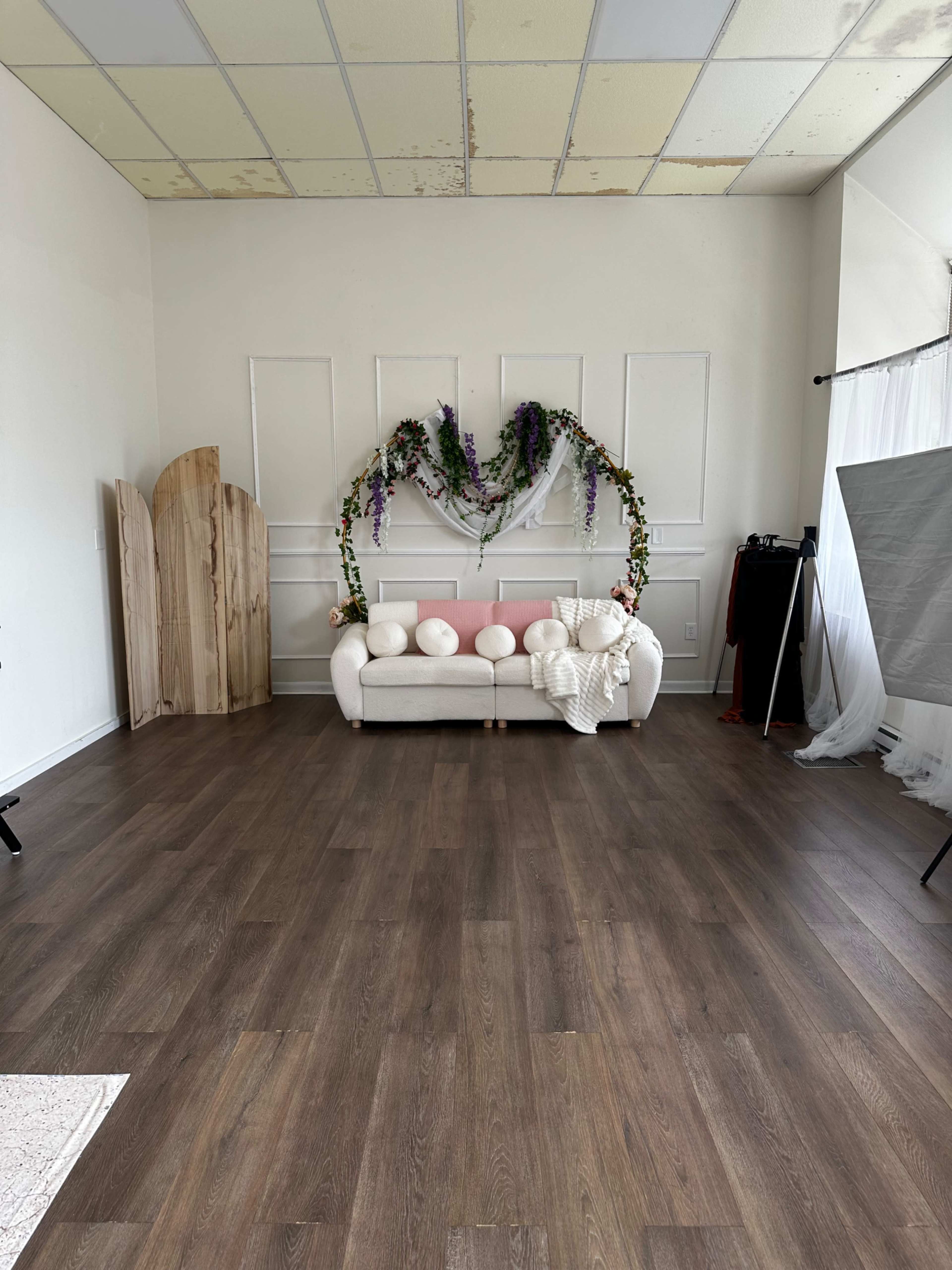 The image shows a spacious room with a white couch adorned with pink pillows, a decorative floral arch above it, and wooden screens in the background.