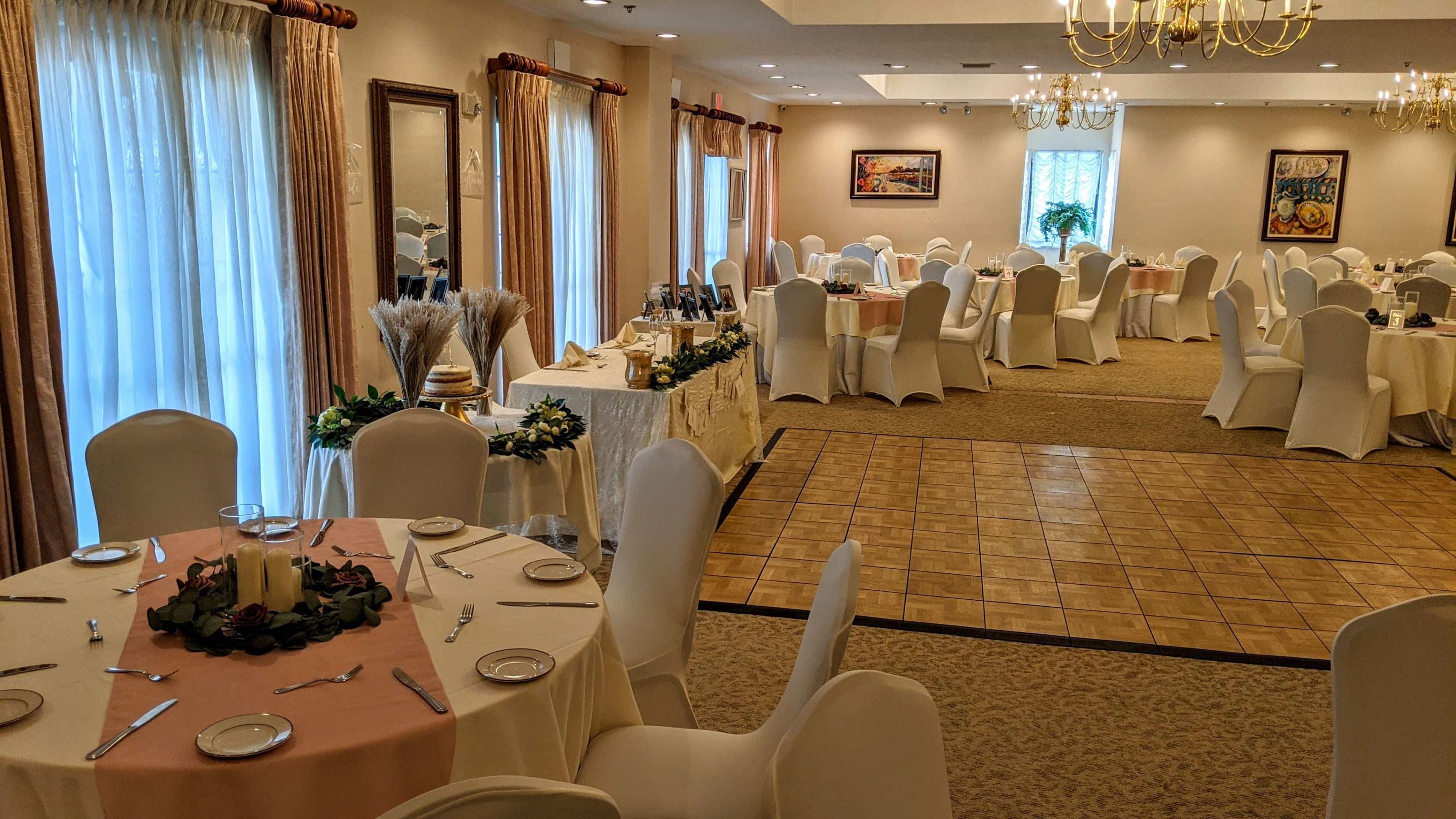 A banquet hall with neatly arranged round tables, white chairs, and a wooden dance floor, surrounded by elegant decor and natural light.