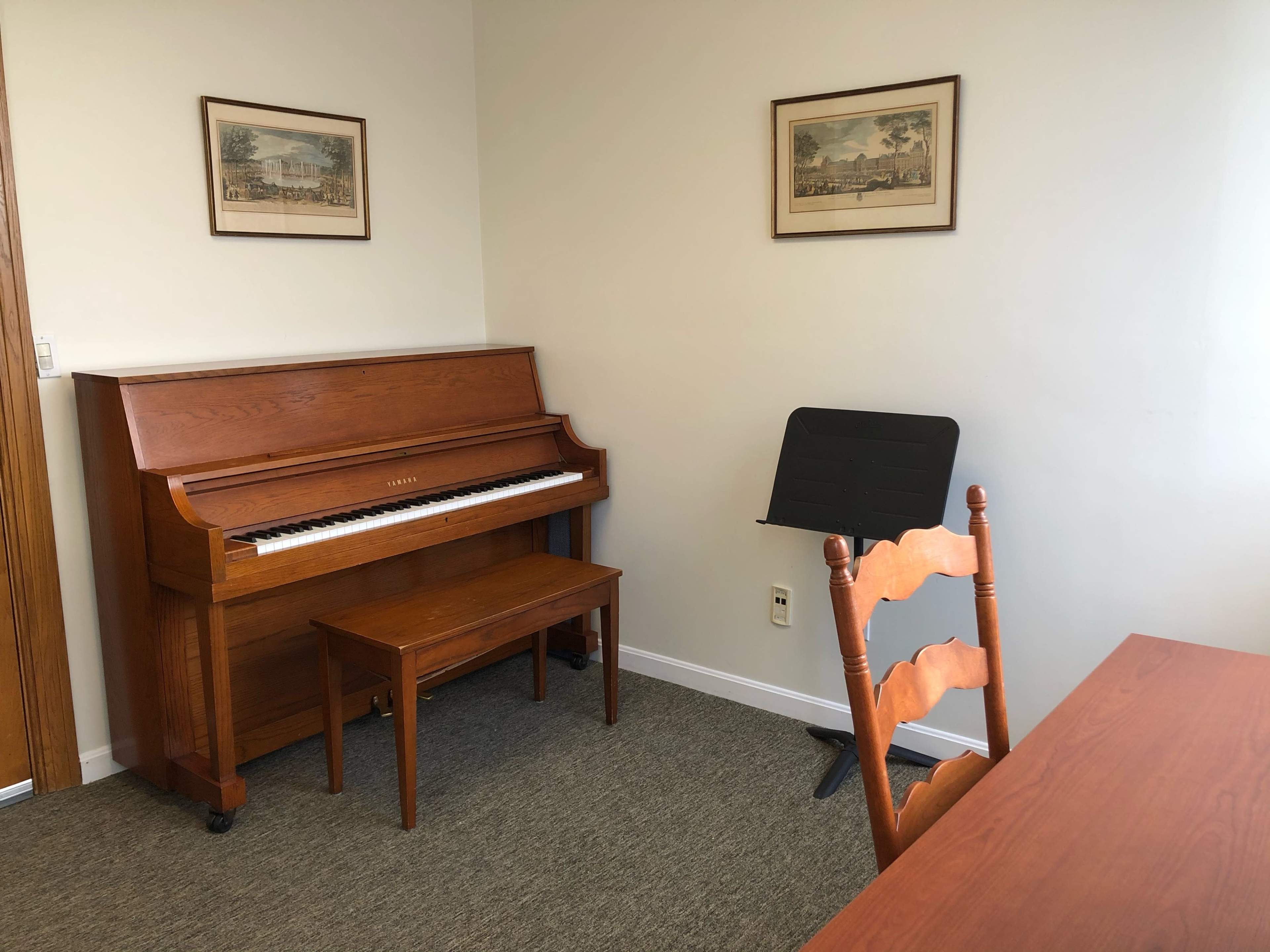 A wooden piano is positioned against a wall in a small, well-lit room featuring a chair and a music stand.