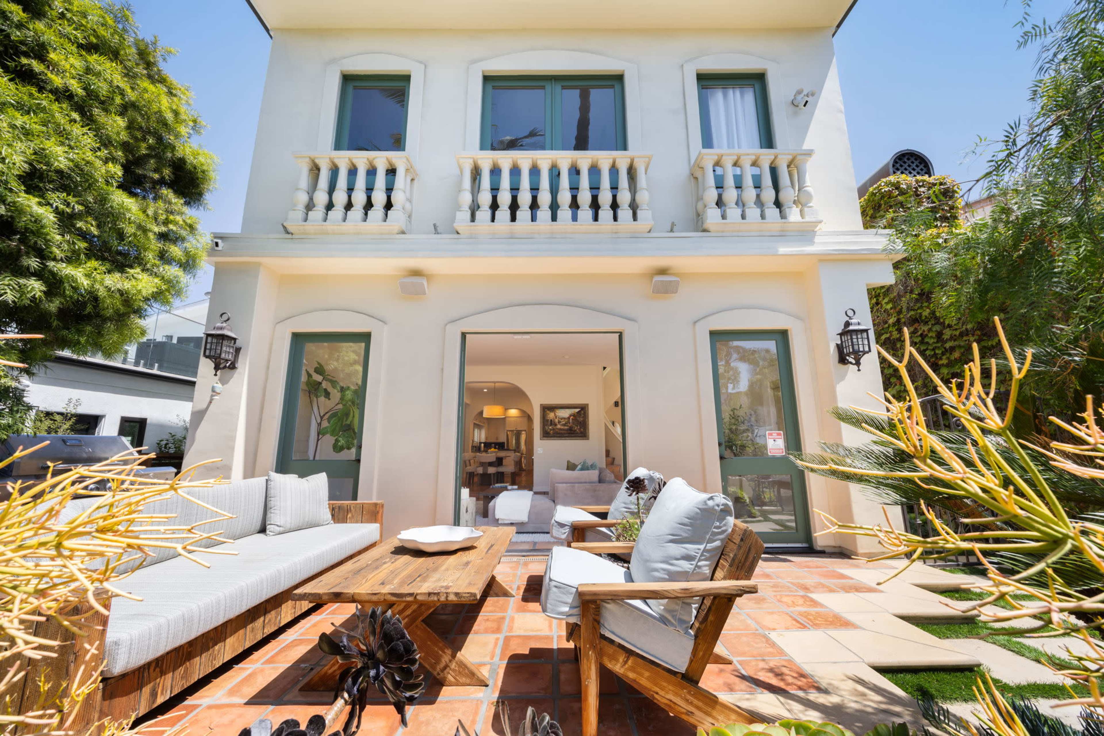 The image shows a modern two-story house with a landscaped patio area featuring outdoor seating and a coffee table.