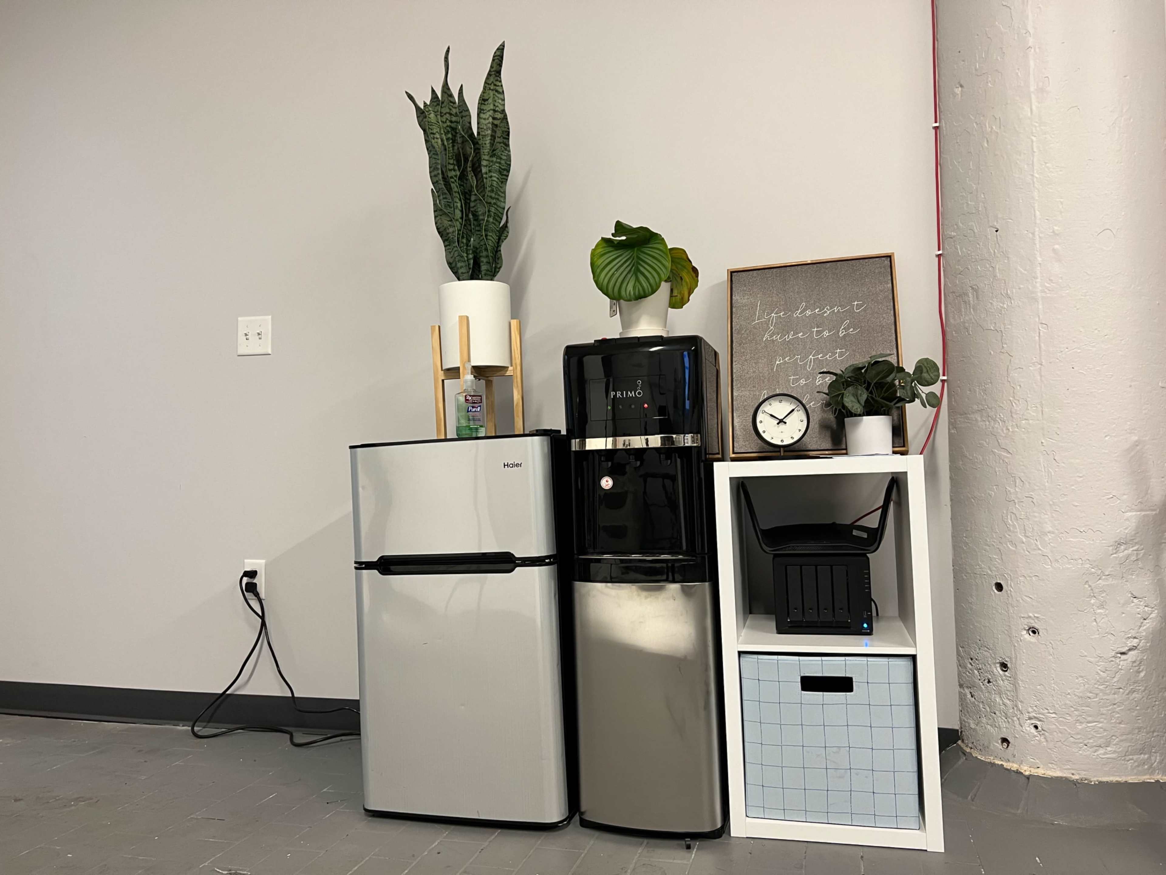 A small kitchen corner with a silver mini fridge, a black water dispenser, and a white shelf featuring a potted plant, clock, and storage box against a gray wall.