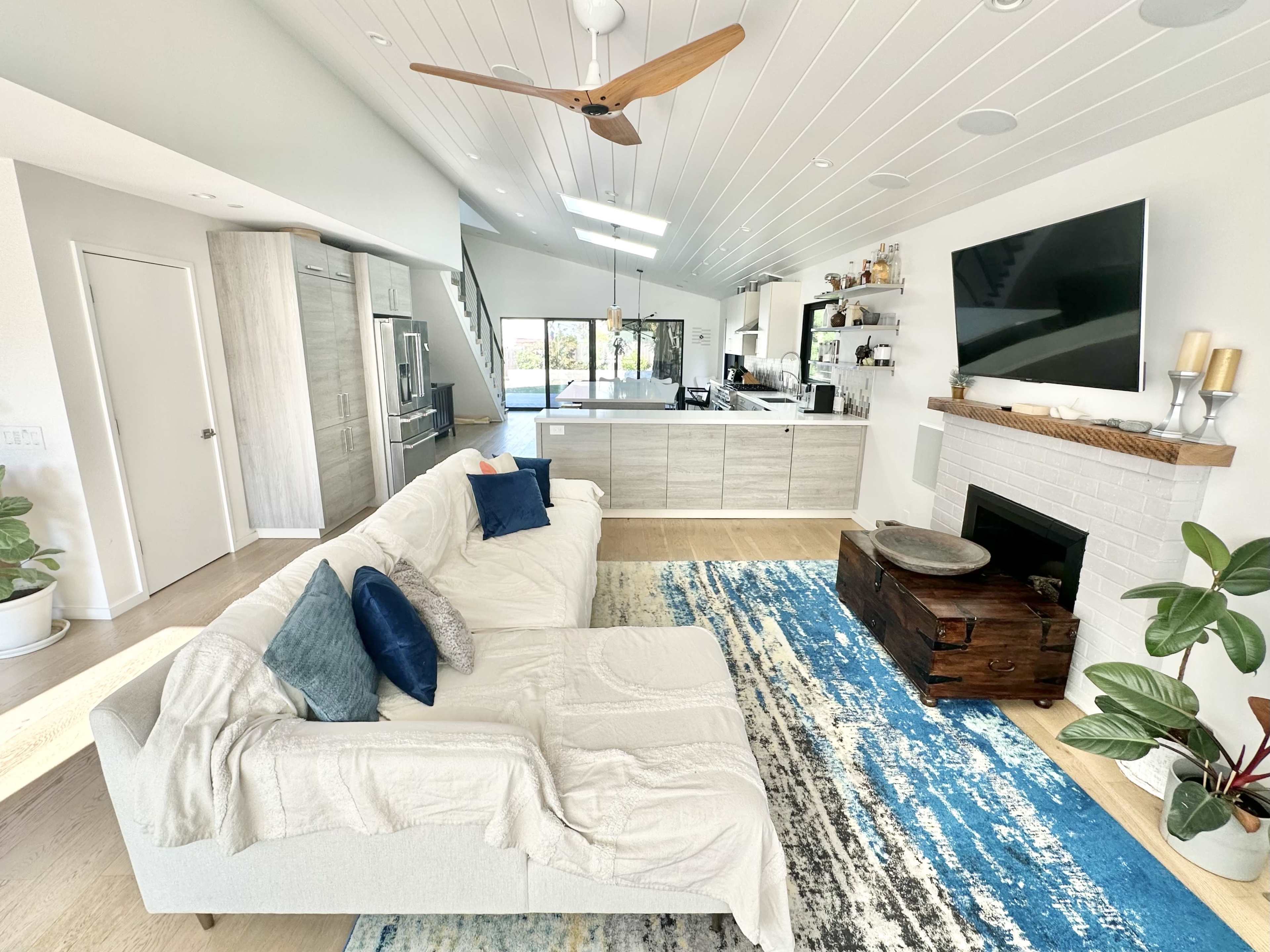 A modern living room featuring a light-colored sectional sofa, a wooden coffee table on a vibrant area rug, and a kitchen area visible in the background.