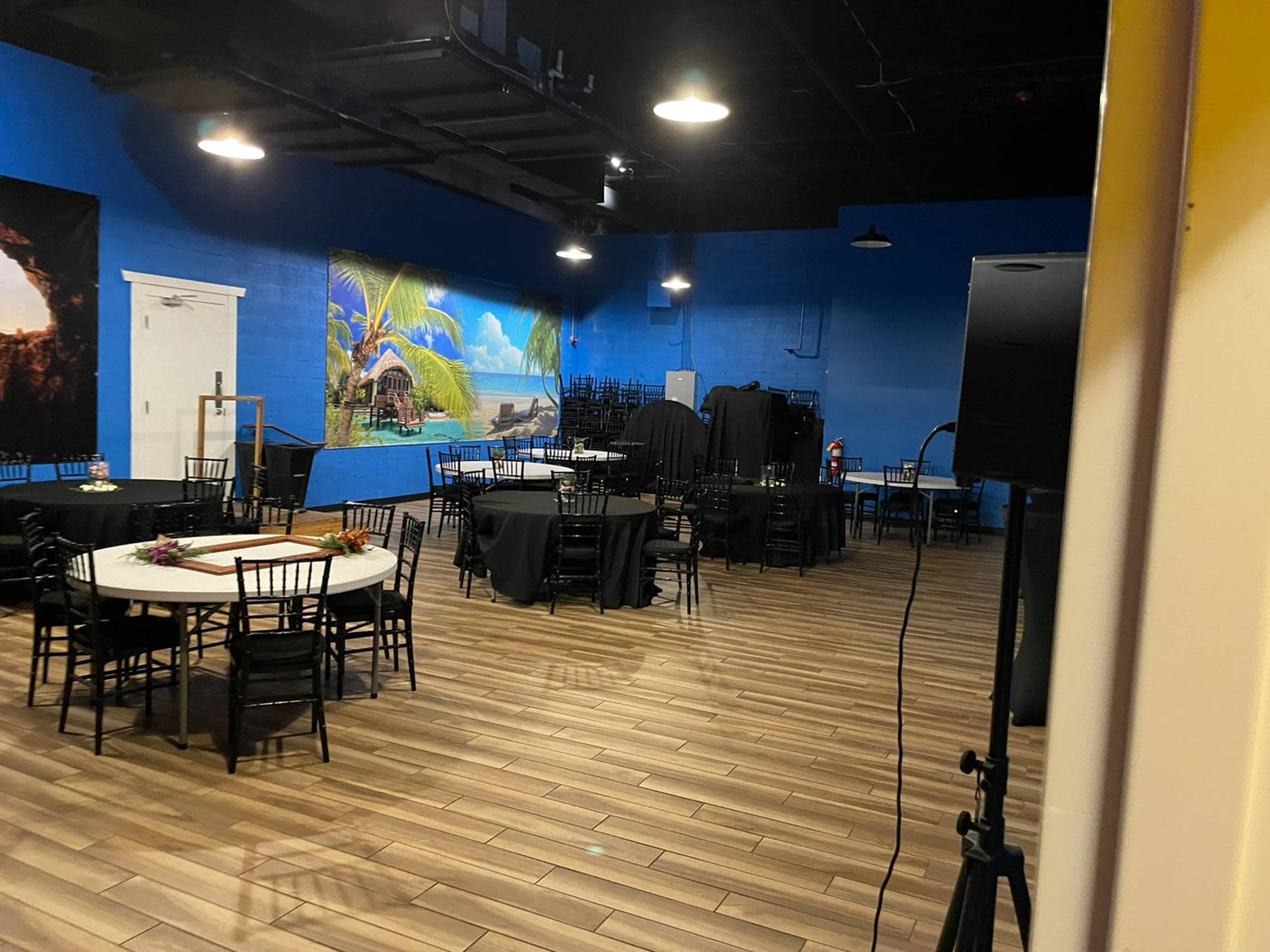 A large event space with black tables and chairs, a blue wall featuring a tropical mural, and some equipment stacked in the corner.