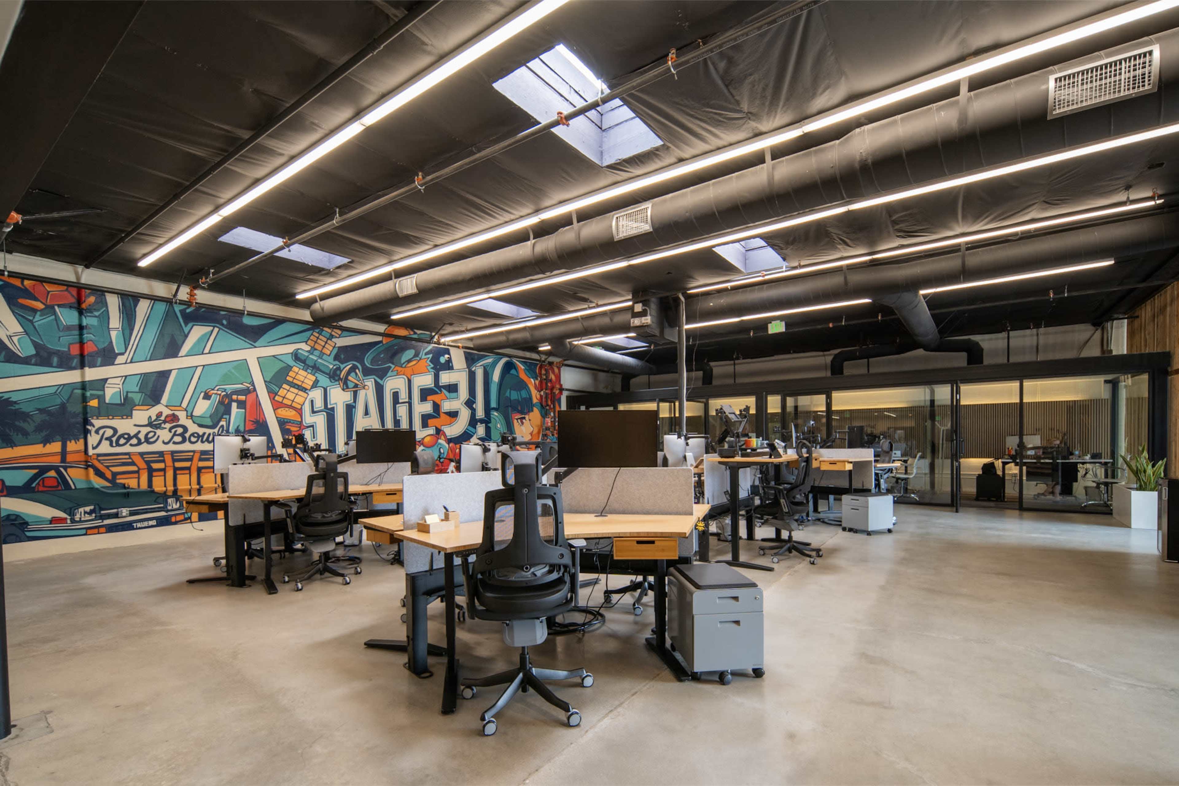 The image shows a modern open office space with several desks, ergonomic chairs, and a large mural on one wall.