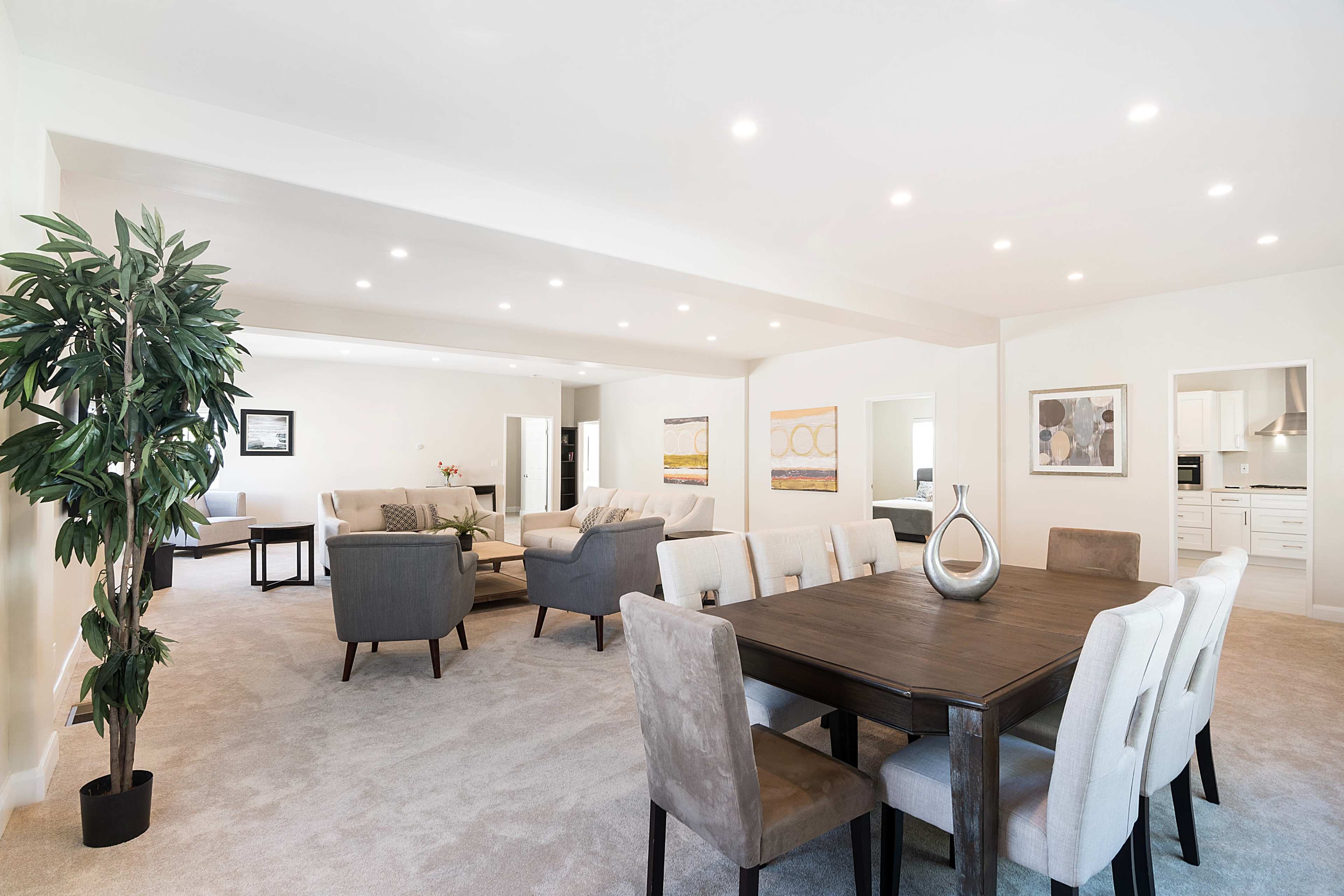 The image shows a modern, open-concept living and dining area with a large dining table, upholstered chairs, and soft carpeting, illuminated by recessed lighting.