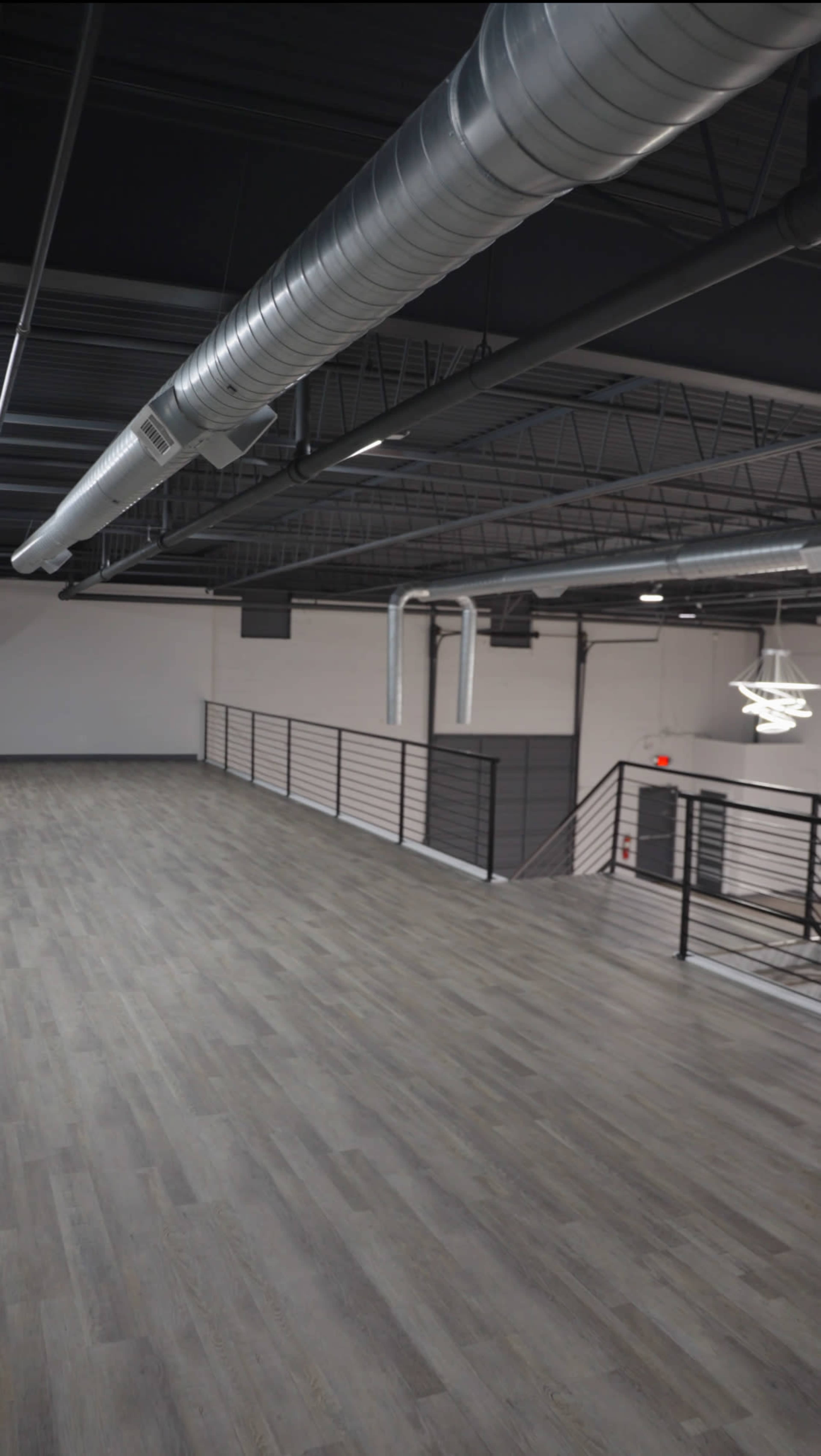 The image shows a spacious, empty room with wooden flooring, high ceilings, metal ductwork, and a railing along a staircase.