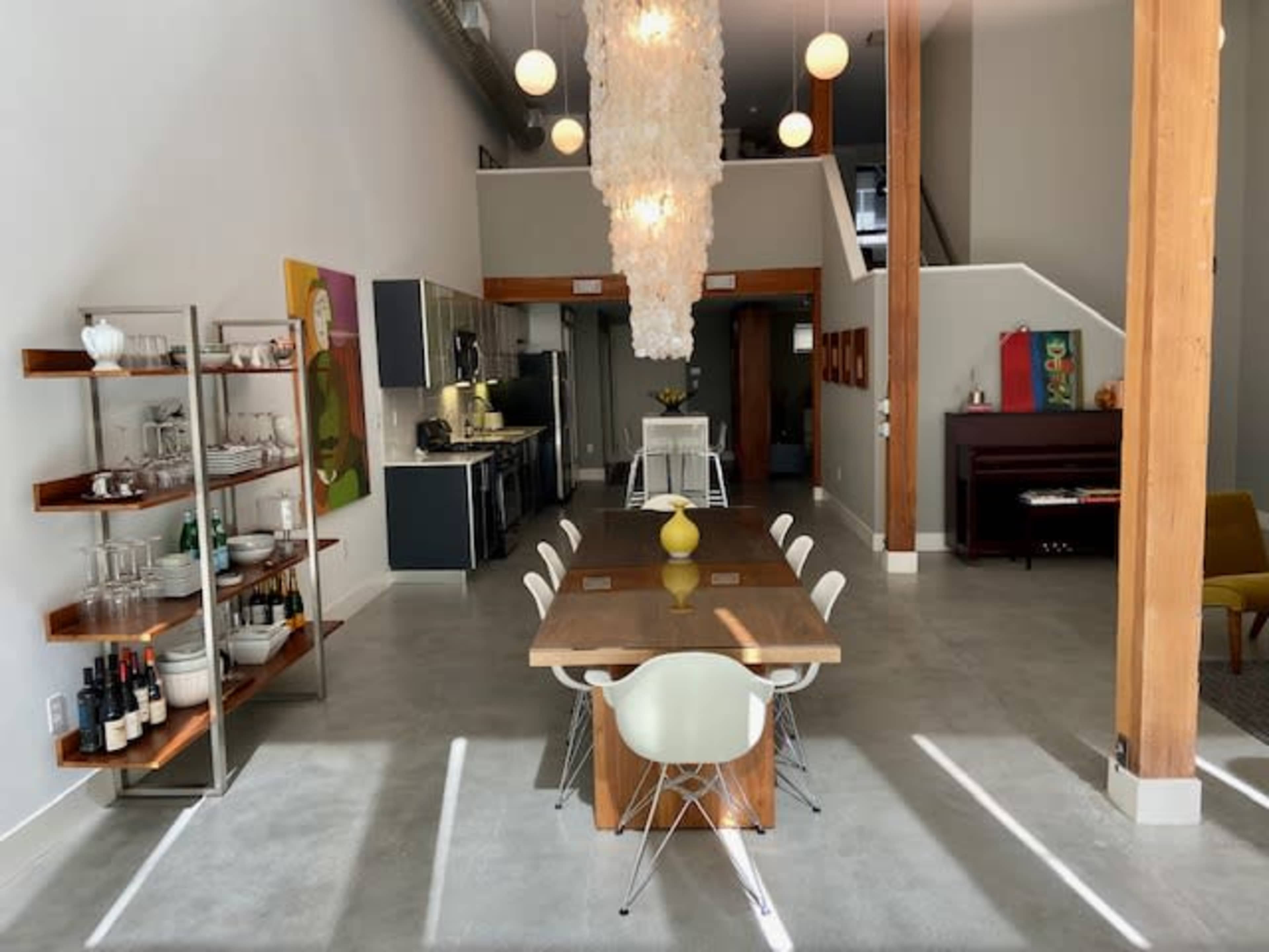 A modern dining area featuring a long wooden table, white chairs, and a large light fixture, with a kitchen and staircase visible in the background.