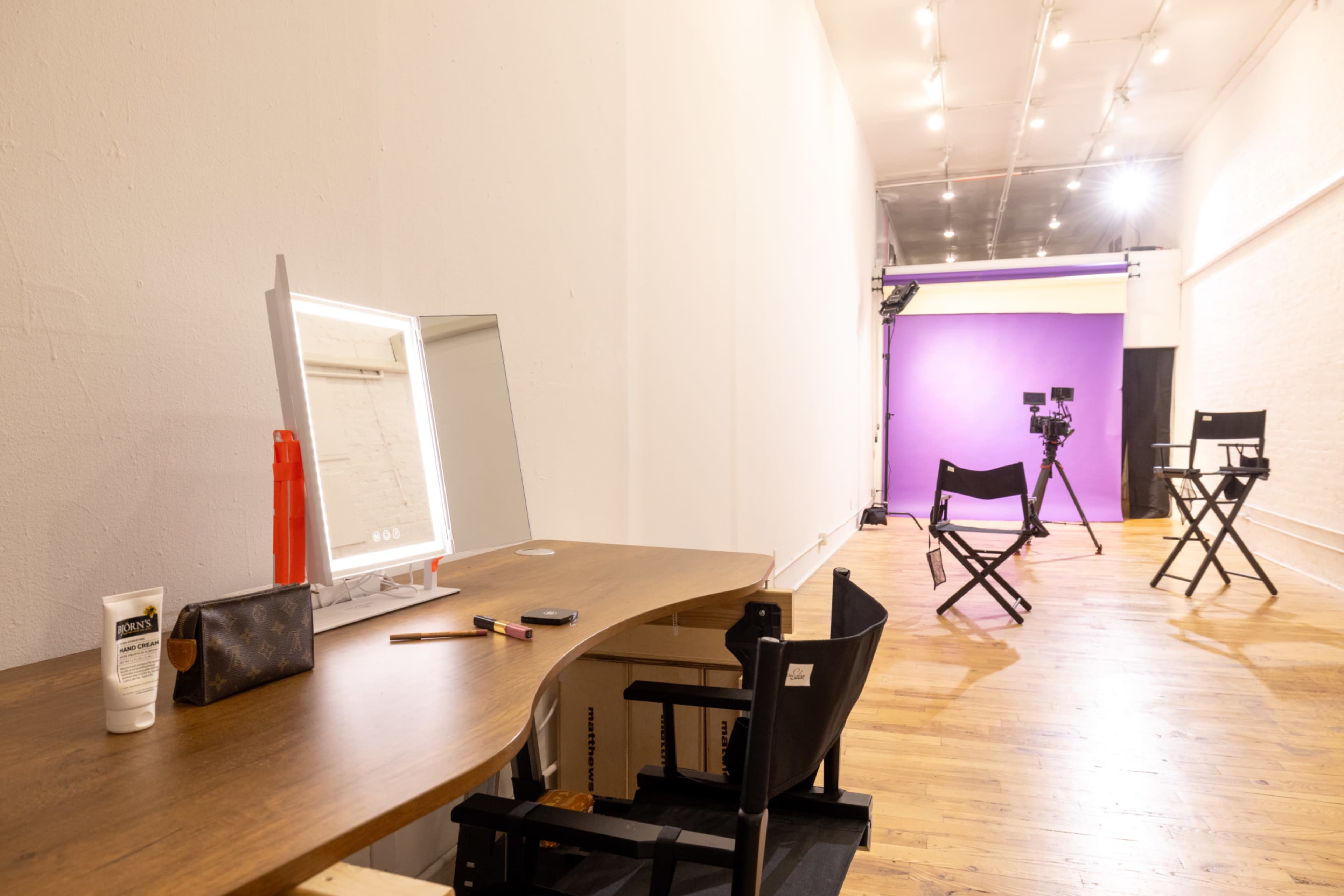 A makeup station with a mirror and cosmetics sits next to a photography area featuring cameras and director's chairs against a purple backdrop.