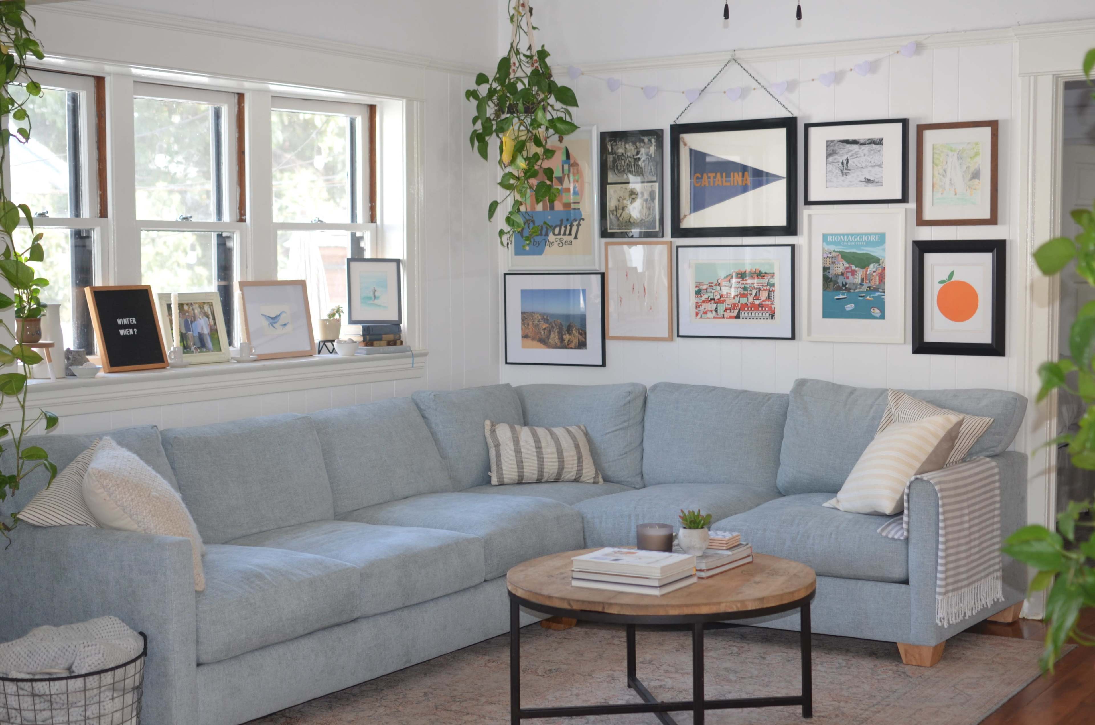 The image shows a cozy, well-decorated living room featuring a blue sectional couch, a round wooden coffee table, and a wall adorned with various framed artworks and photographs.