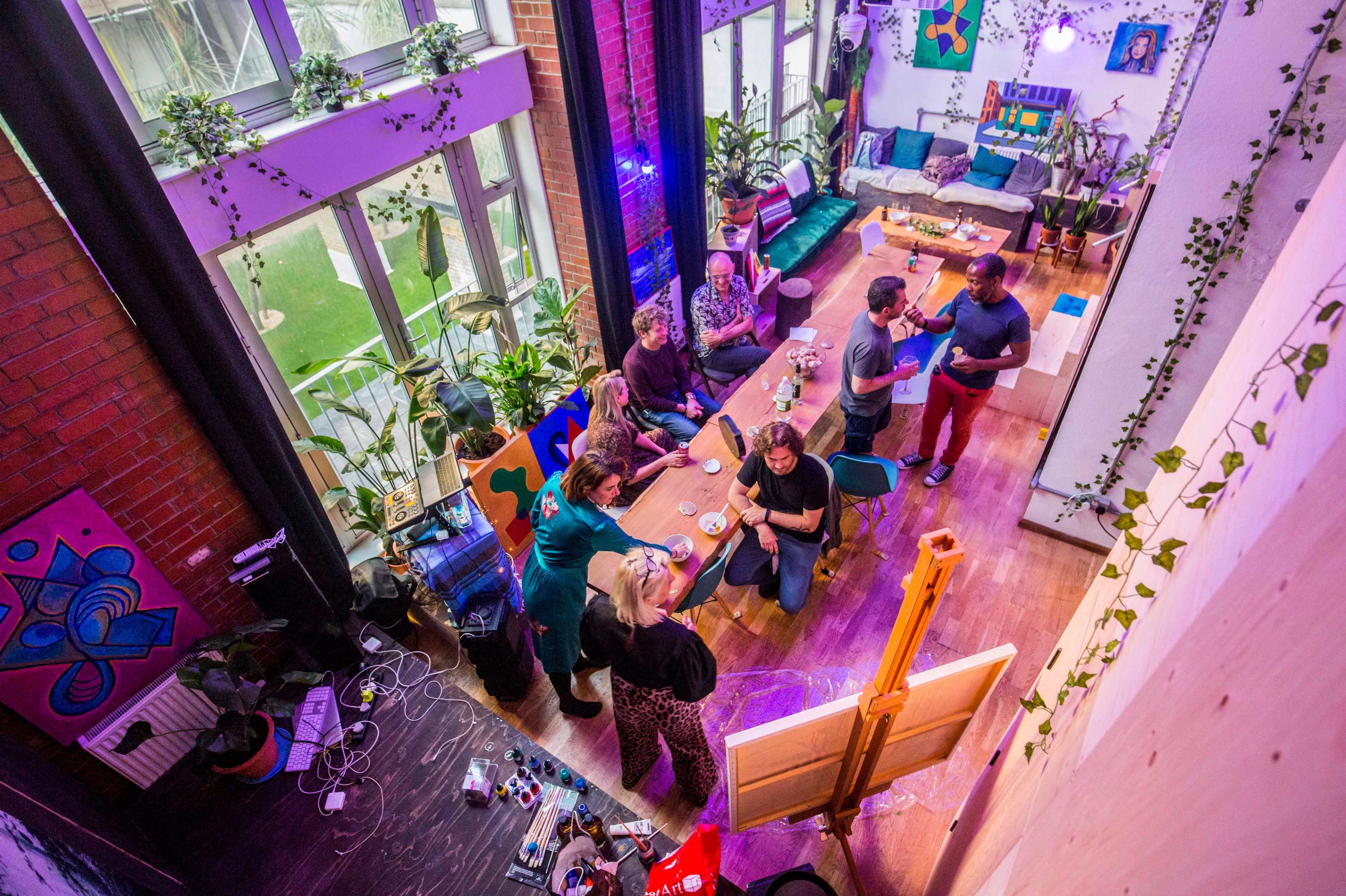 A group of people engages in conversation around a table in a vibrant, plant-filled interior space featuring colorful art and large windows.