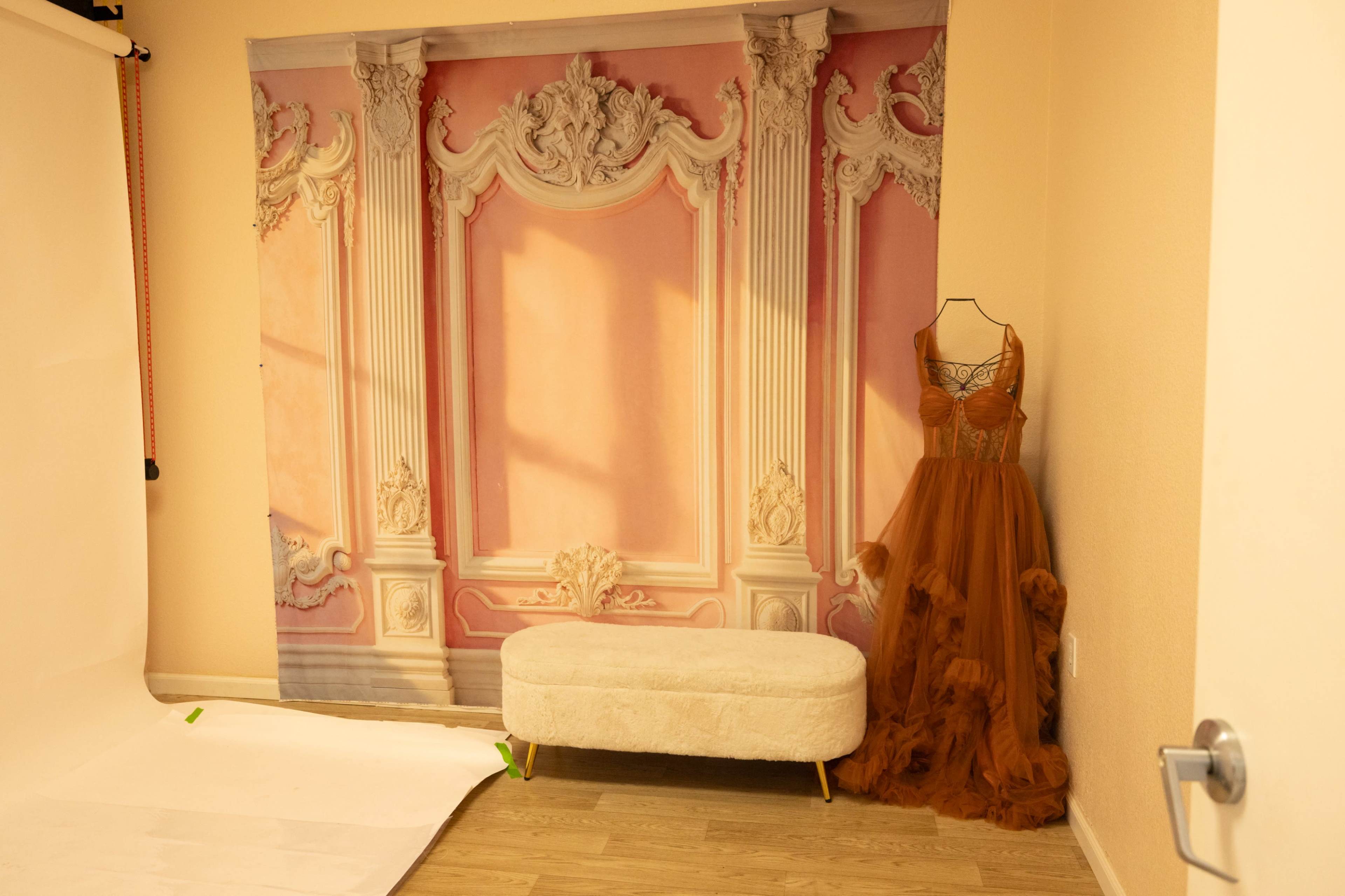 The image shows a photography studio with a decorative backdrop of ornate pink walls and a formal gown displayed on a mannequin beside a small upholstered bench.