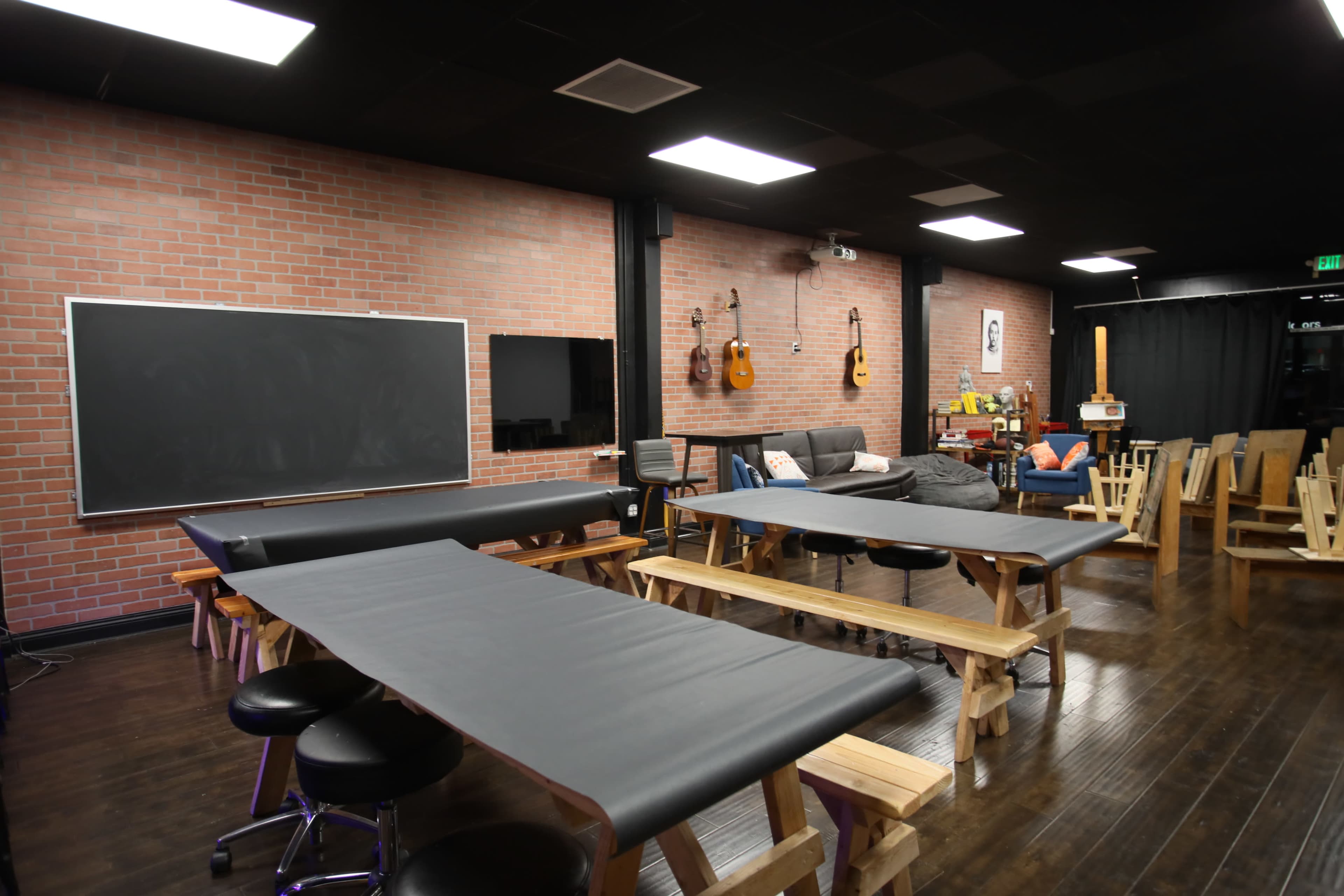 A spacious room with wooden tables and stools, a chalkboard on the wall, and acoustic guitars hanging in the background.