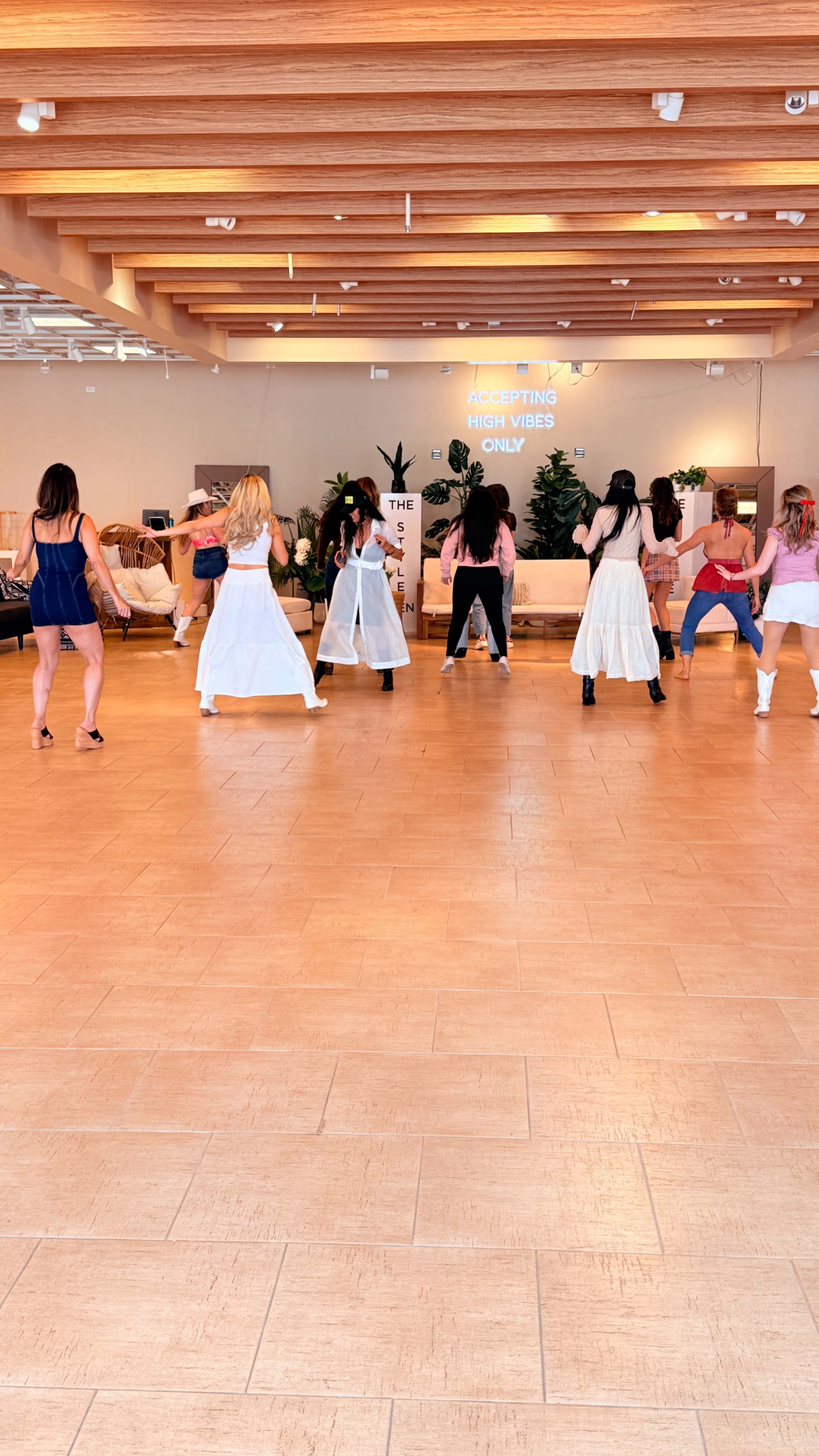 A group of women are dancing in a brightly lit studio with wooden beams and a large screen displaying text in the background.