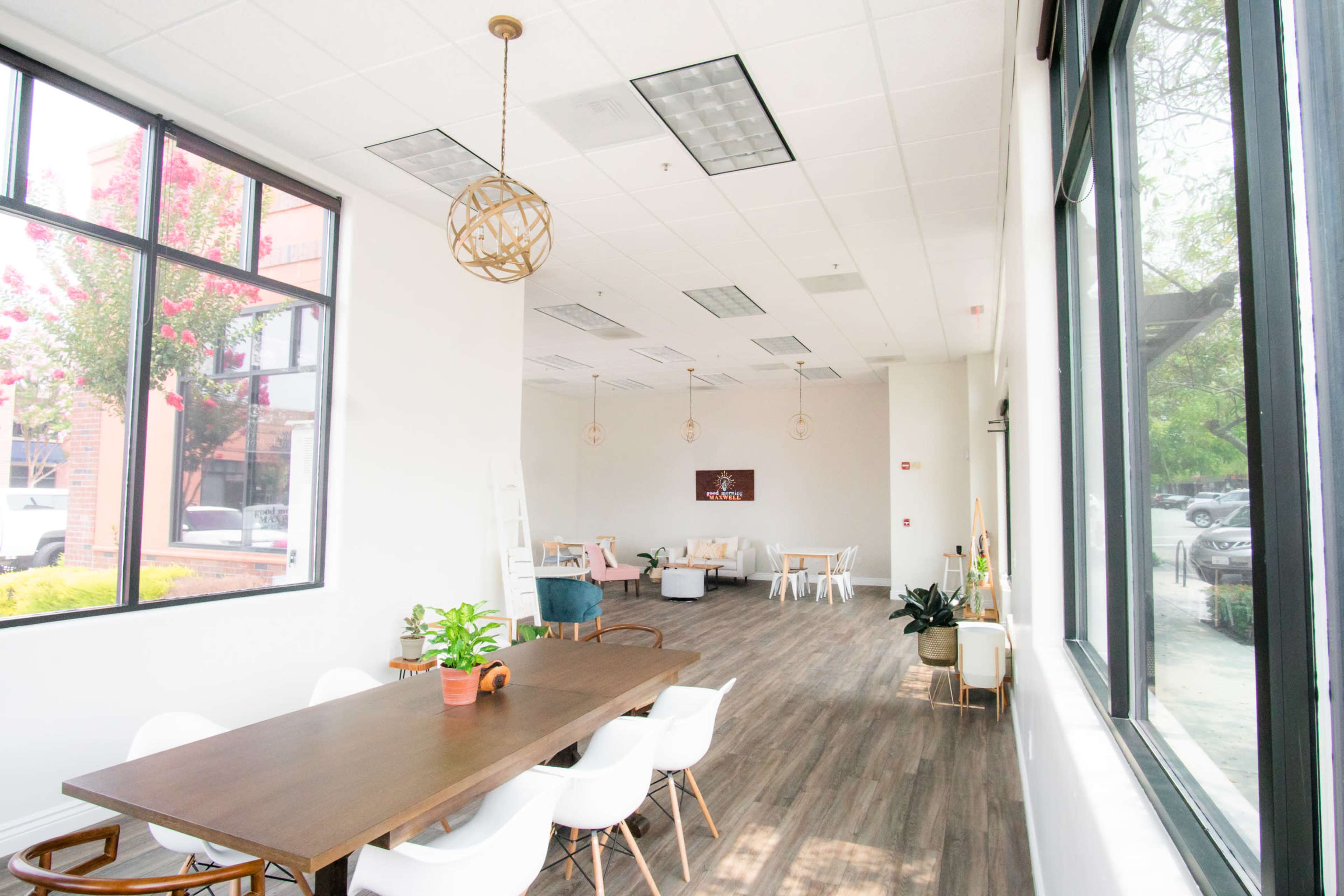 A spacious, well-lit room featuring a long wooden table, white chairs, and large windows with greenery visible outside.