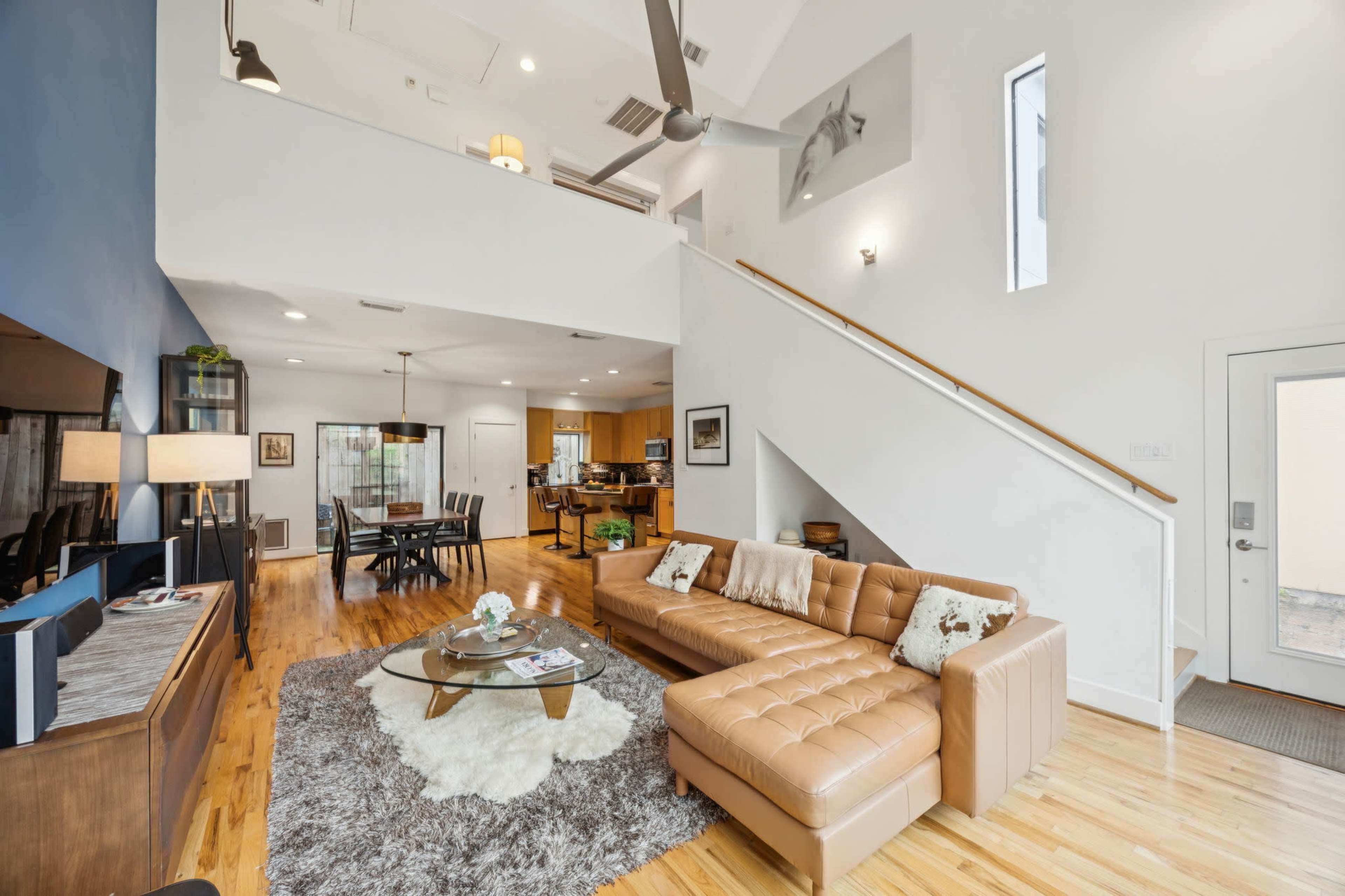 The image shows a modern living room with a tan sectional sofa, a round coffee table on a rug, and kitchen and dining areas visible in the background.