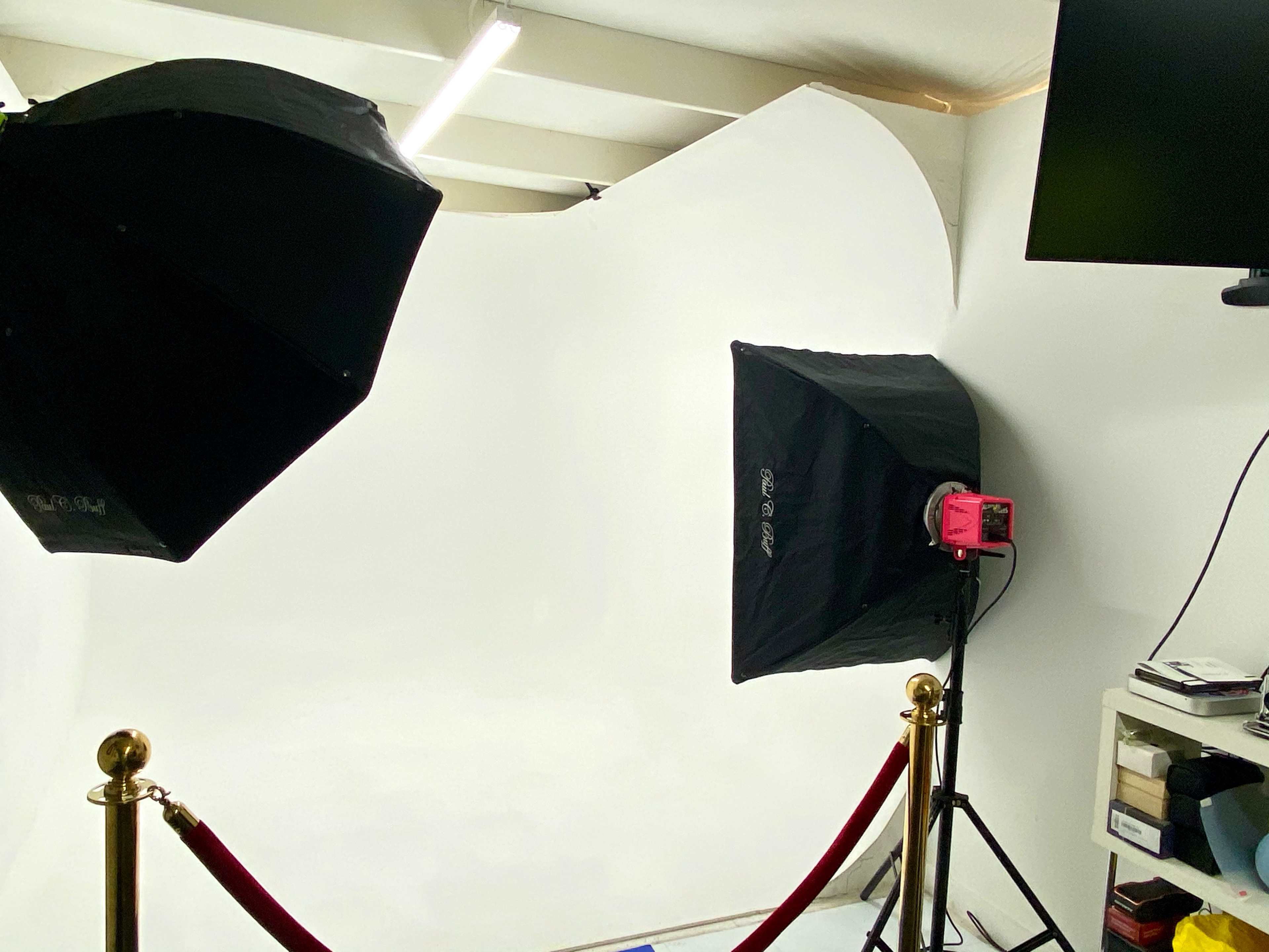 A photography studio setup with two softbox lights, a white backdrop, and a section of red velvet ropes.