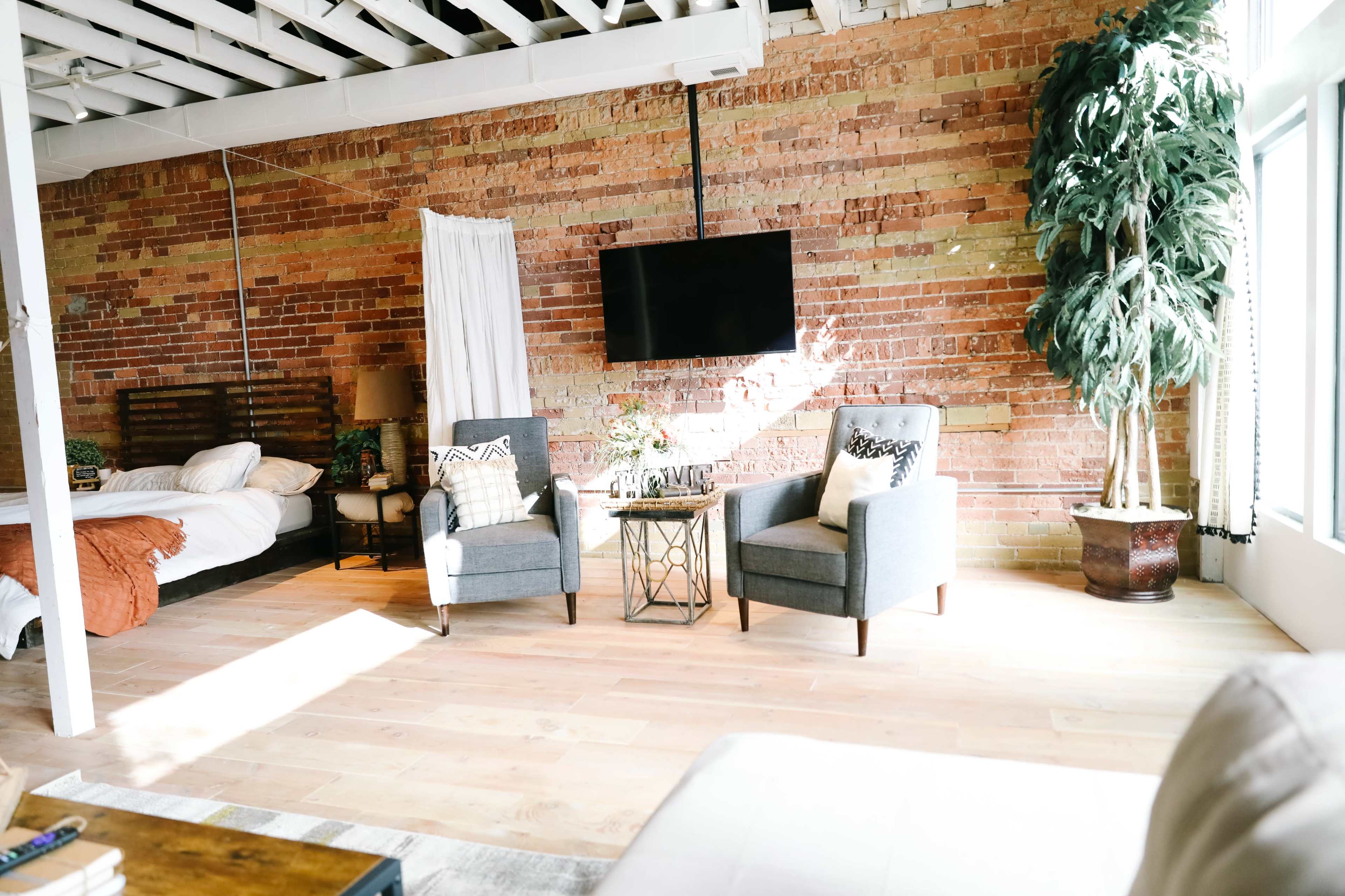 A modern living space with exposed brick walls, two gray chairs and a small table, a bed in the background, and a large potted plant.