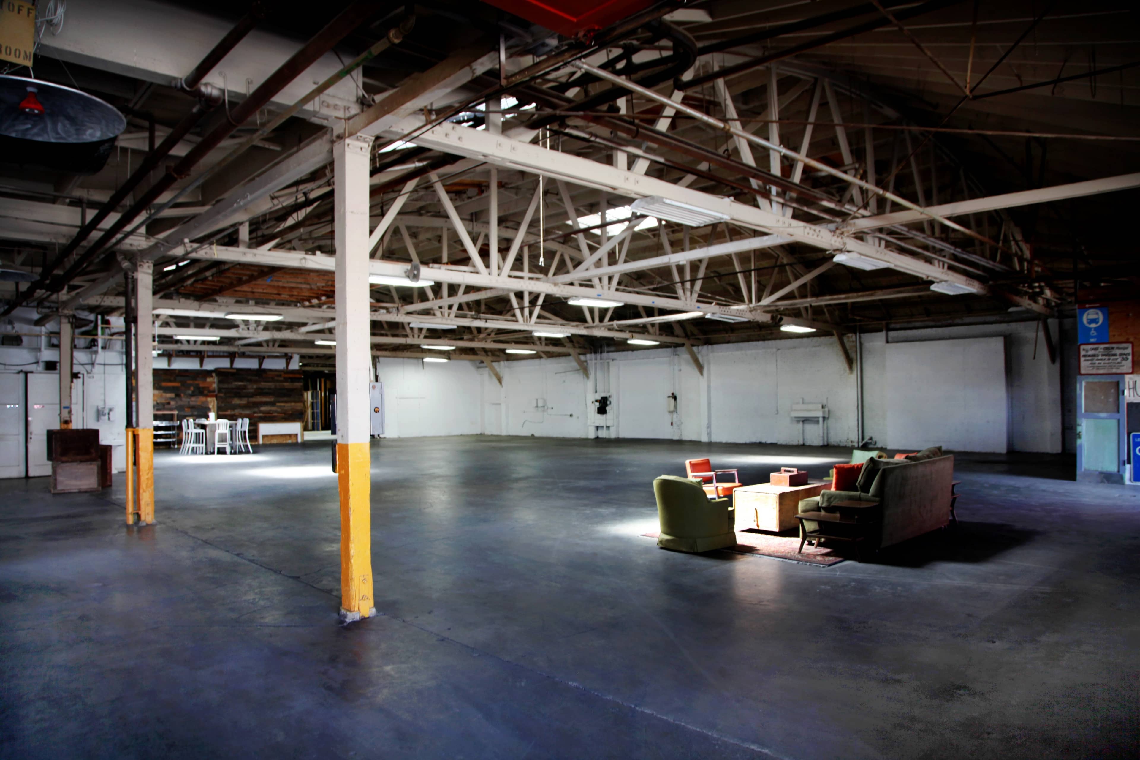 The image shows a spacious, industrial-style interior with exposed beams, concrete floors, and a seating area featuring a sofa and chairs in one corner.