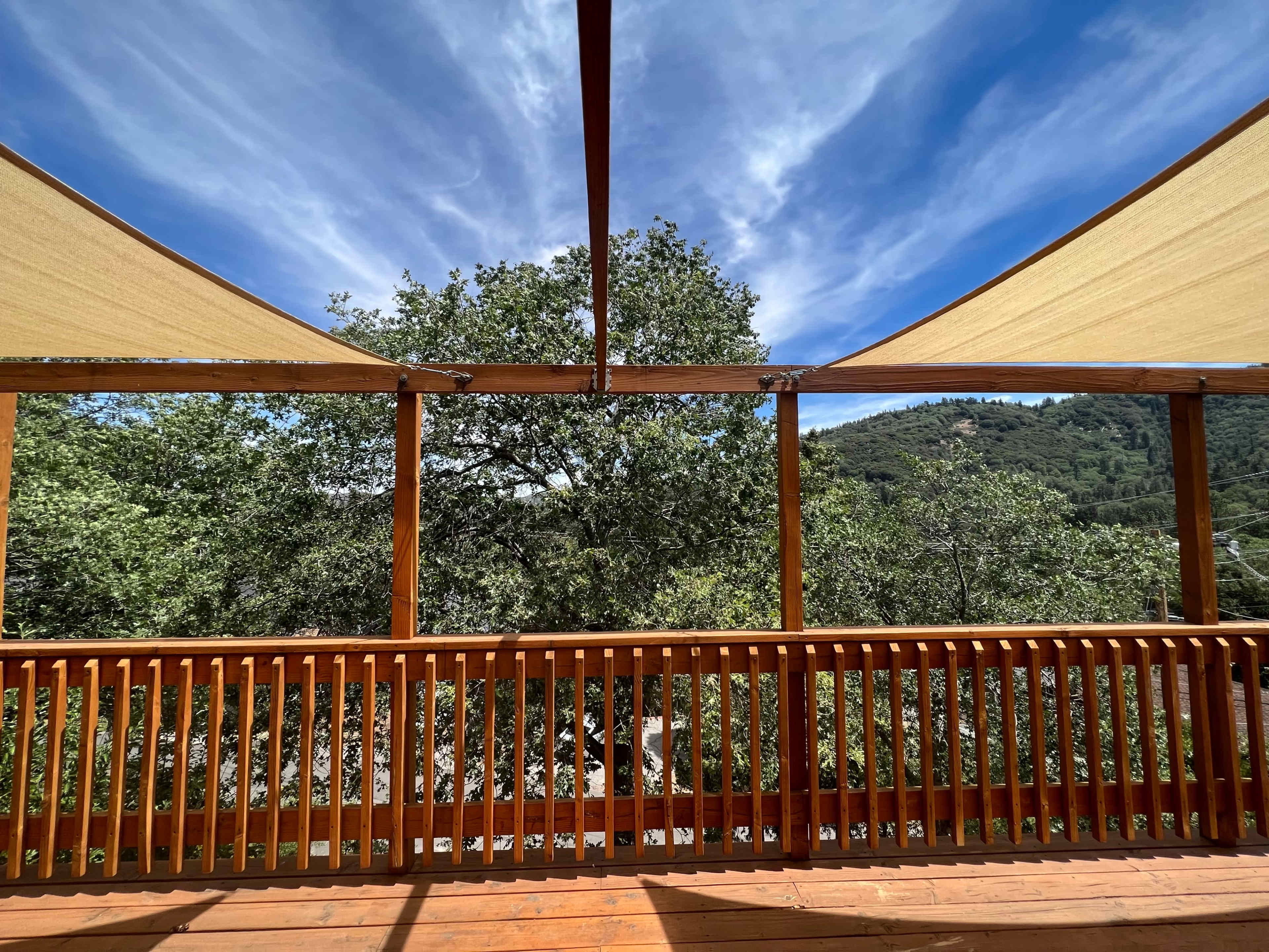 A wooden deck with a railing, overlooking trees and a mountainous landscape under a partly cloudy sky.