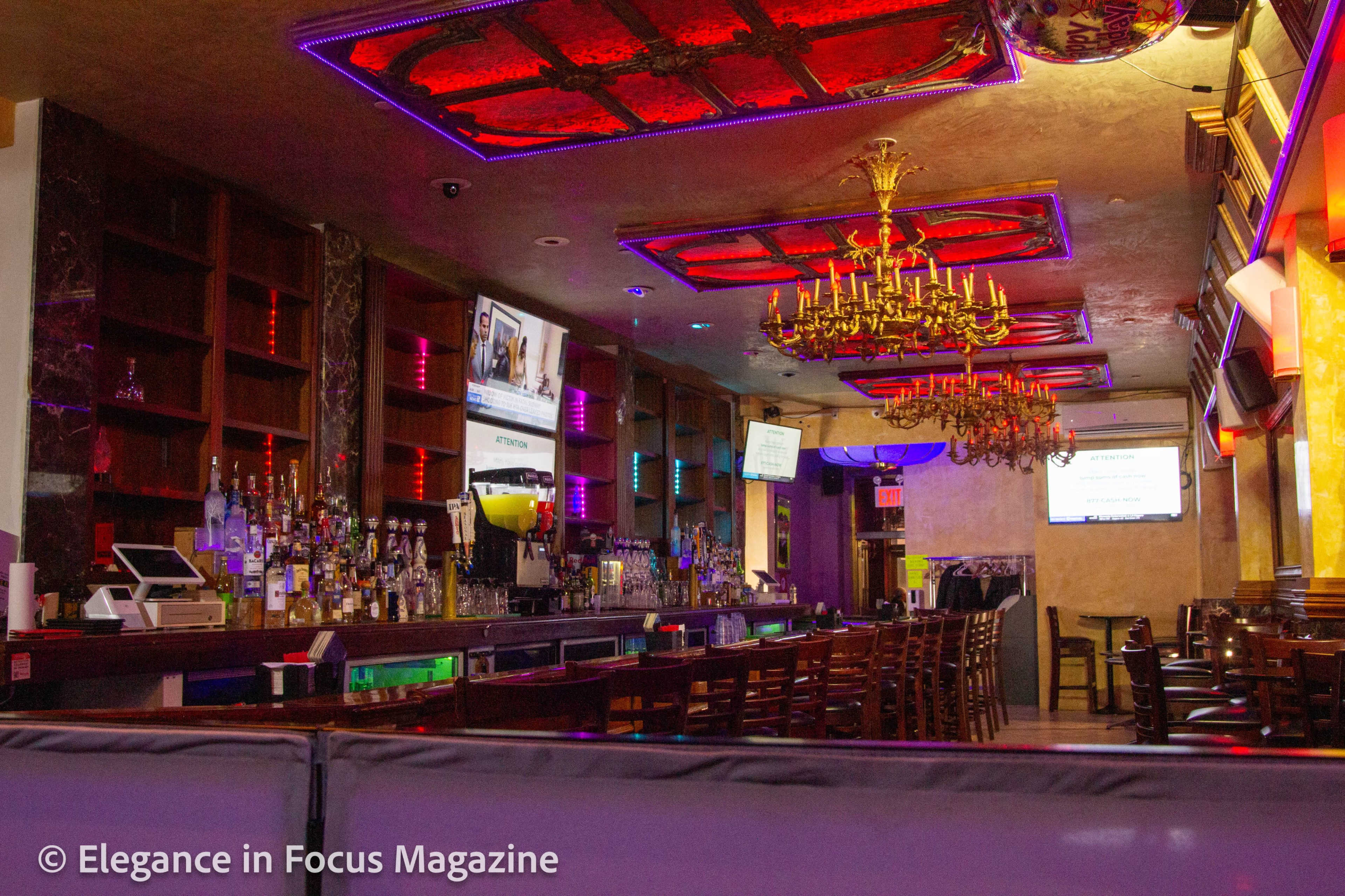 An interior view of a bar with a chandelier, multiple screens, and a well-stocked bar counter.