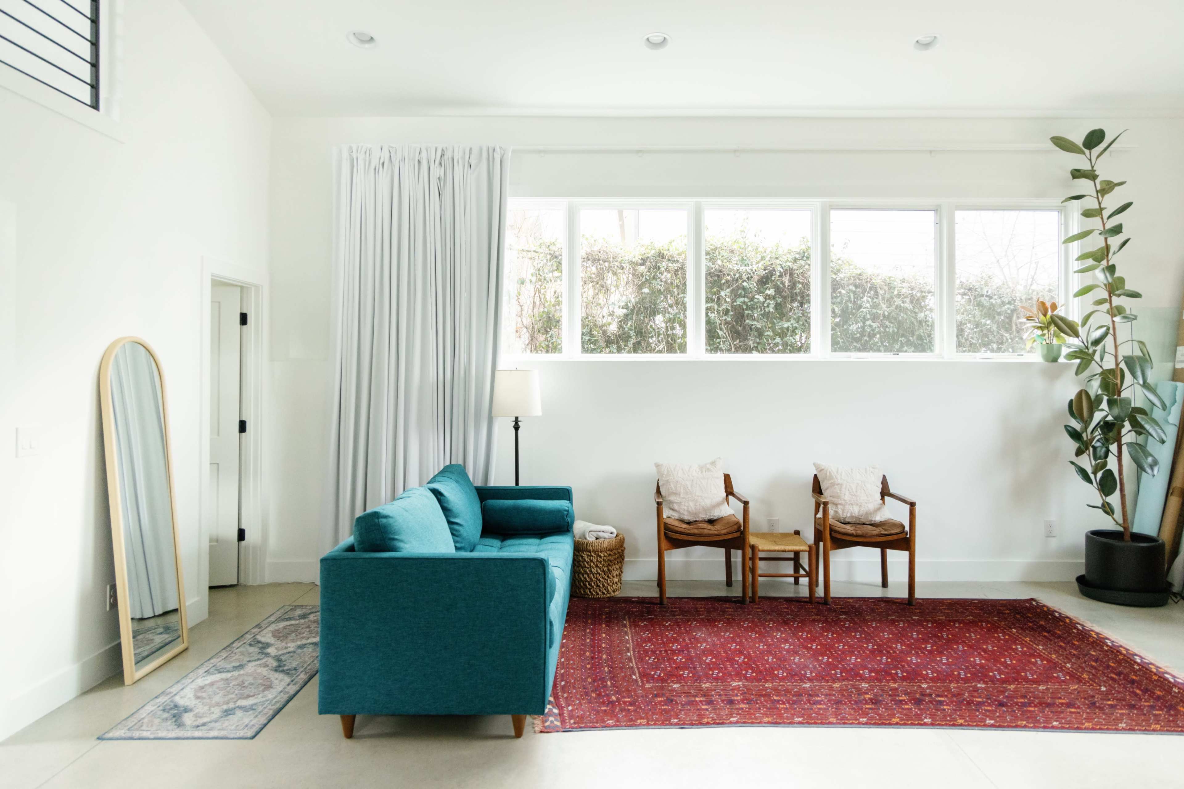 The image shows a bright living room with a blue sofa, two wooden chairs, a large mirror, a lamp, and a red area rug against a white wall with large windows.