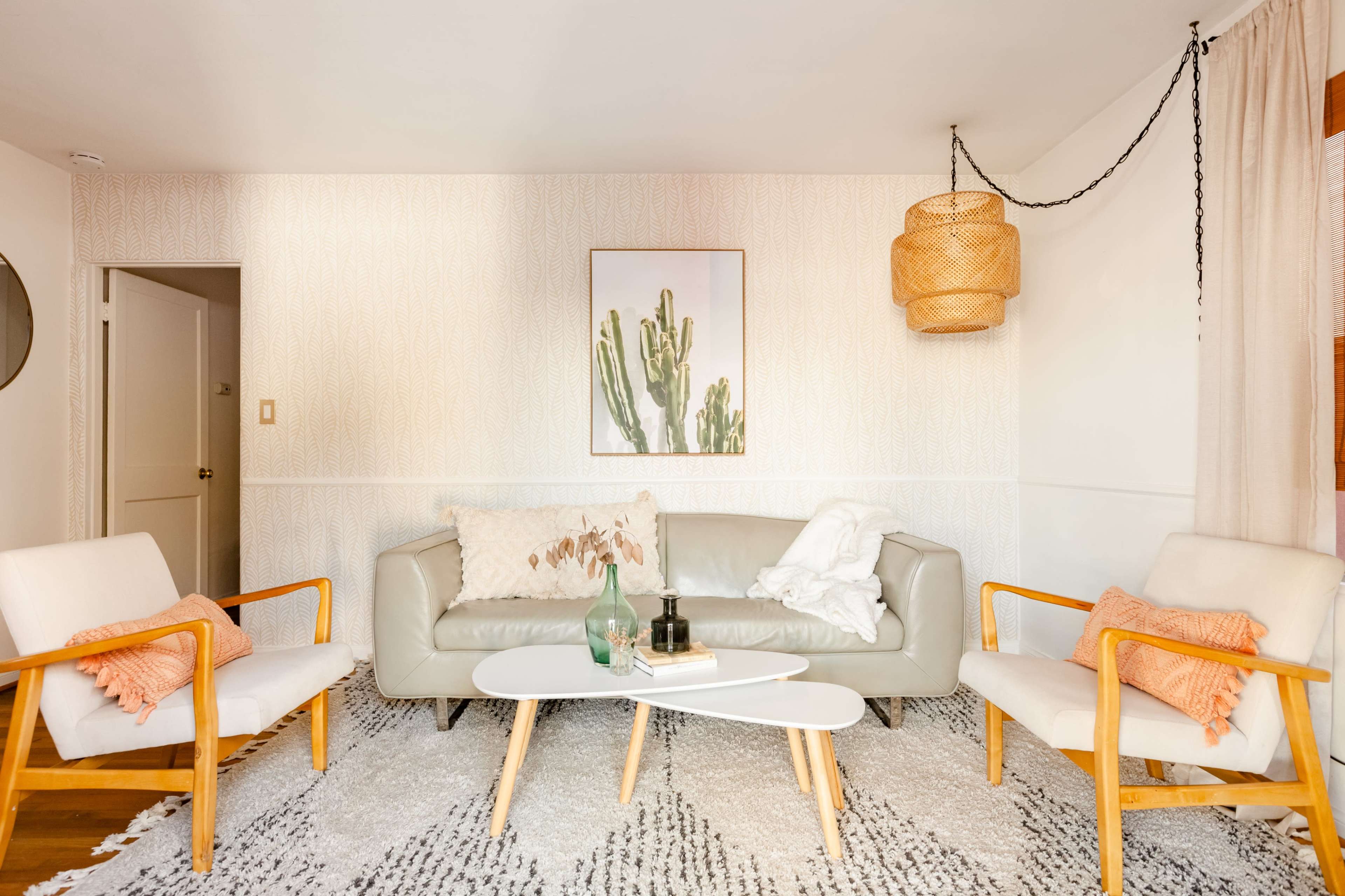 The living room features a light-colored sofa flanked by two wooden chairs, a coffee table, and a wall adorned with a cactus print and a woven pendant light.