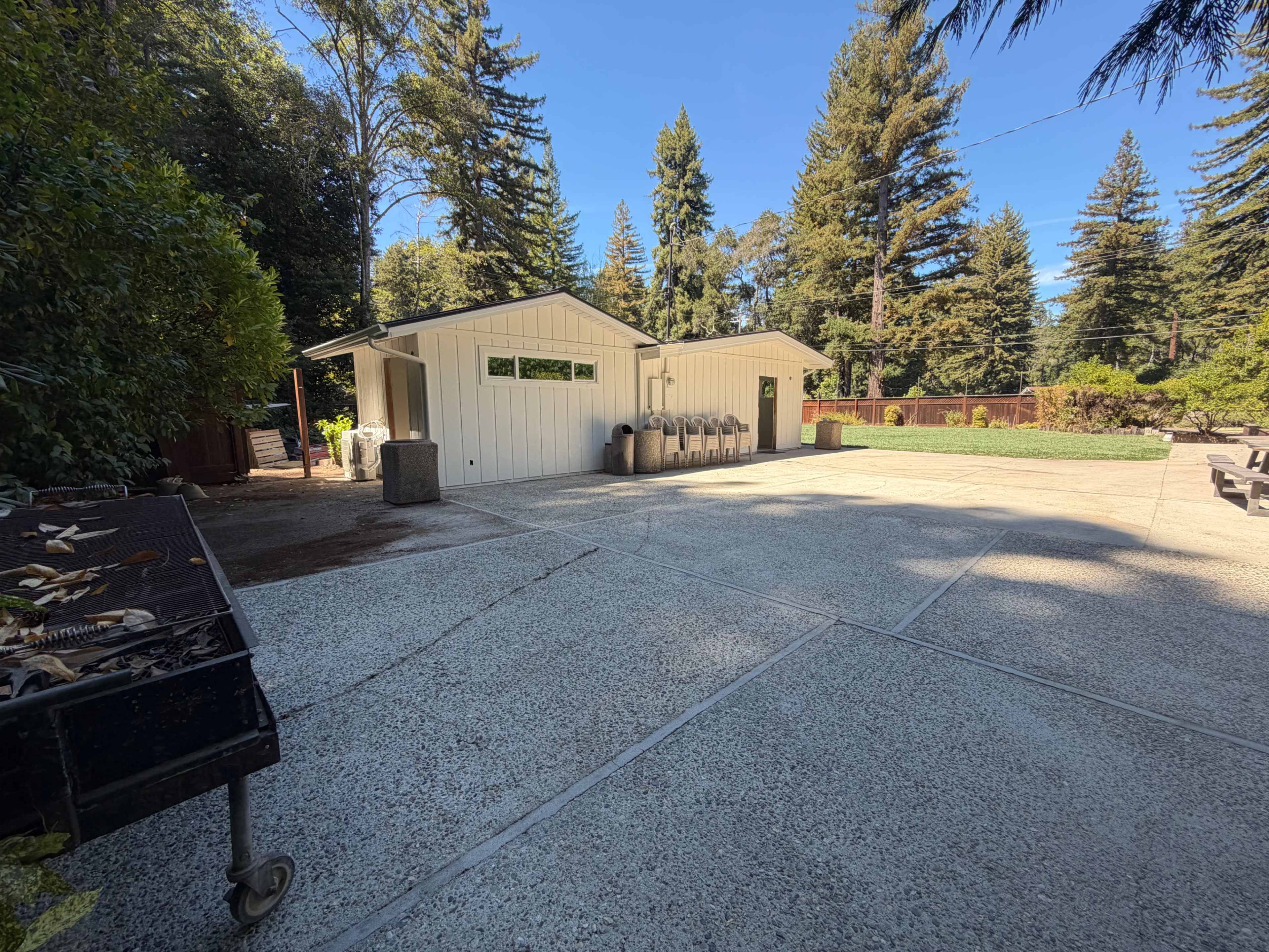 The image shows a concrete outdoor area with two white buildings and tall trees in the background.