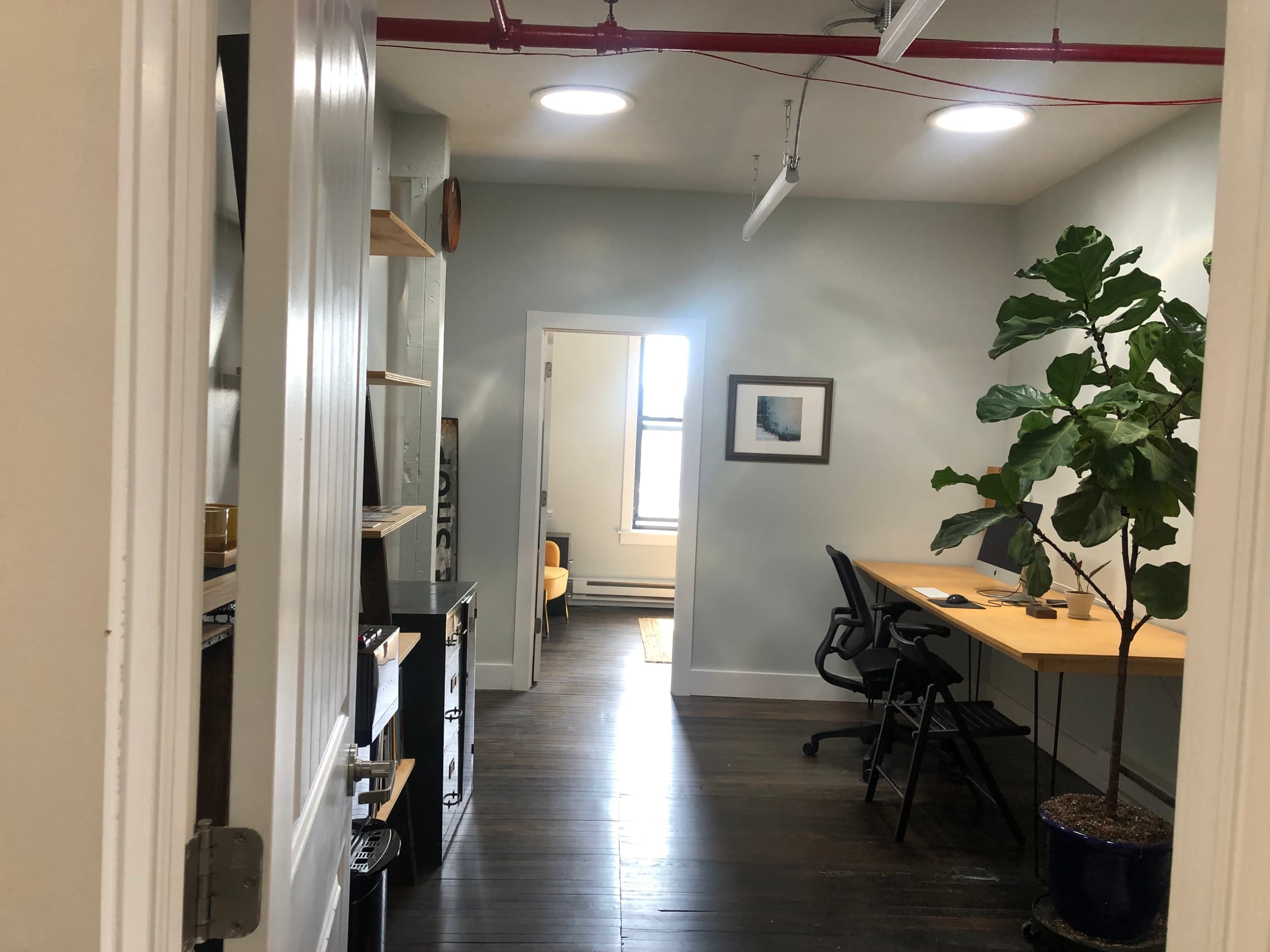 The image shows a hallway leading into a well-lit office space with a desk, a chair, and a potted plant.