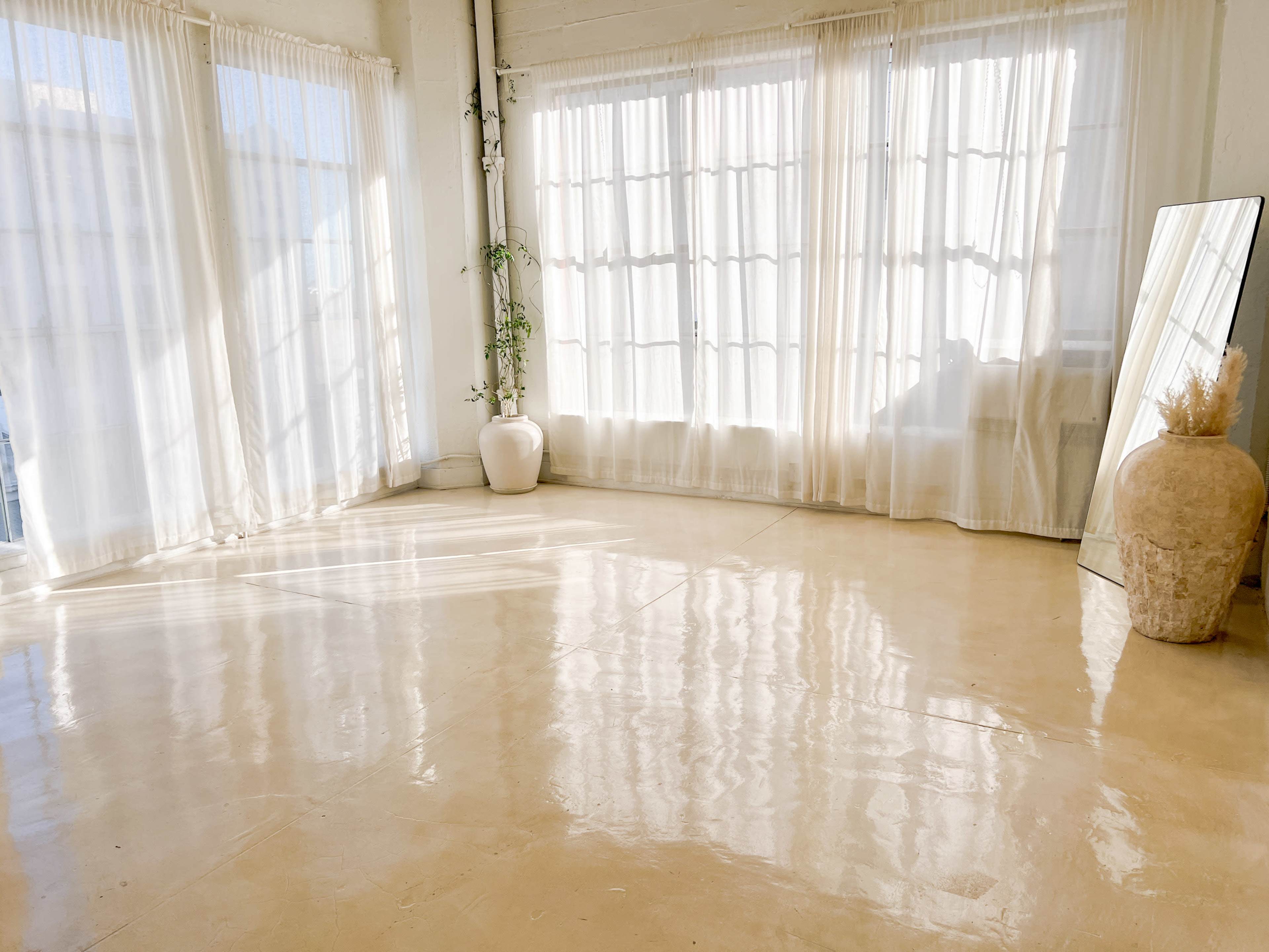 A spacious room with large windows, sheer curtains, and a polished floor, featuring a potted plant and a decorative vase.