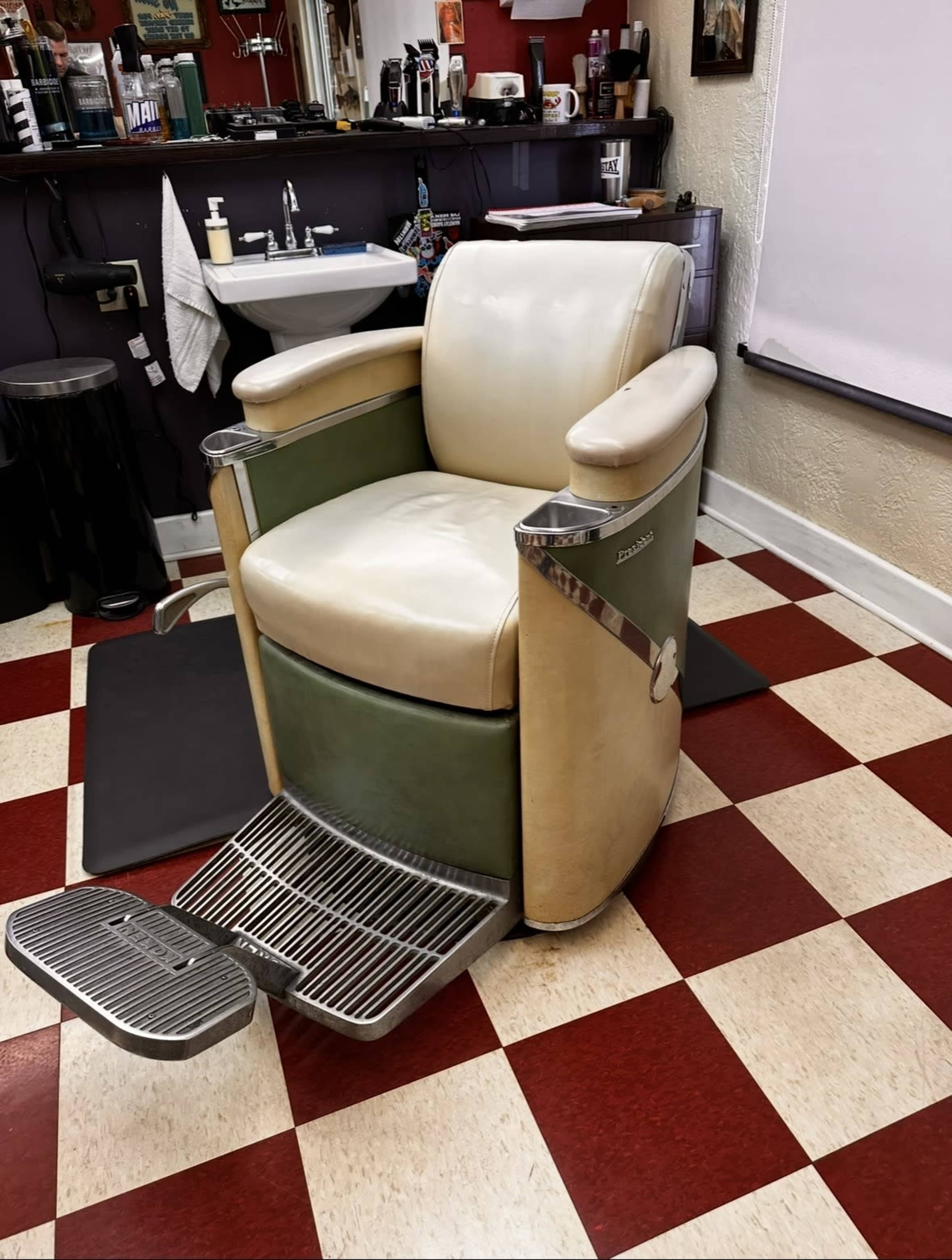 The image shows a vintage barber chair situated on a checkered floor of red and cream tiles in a barber shop.