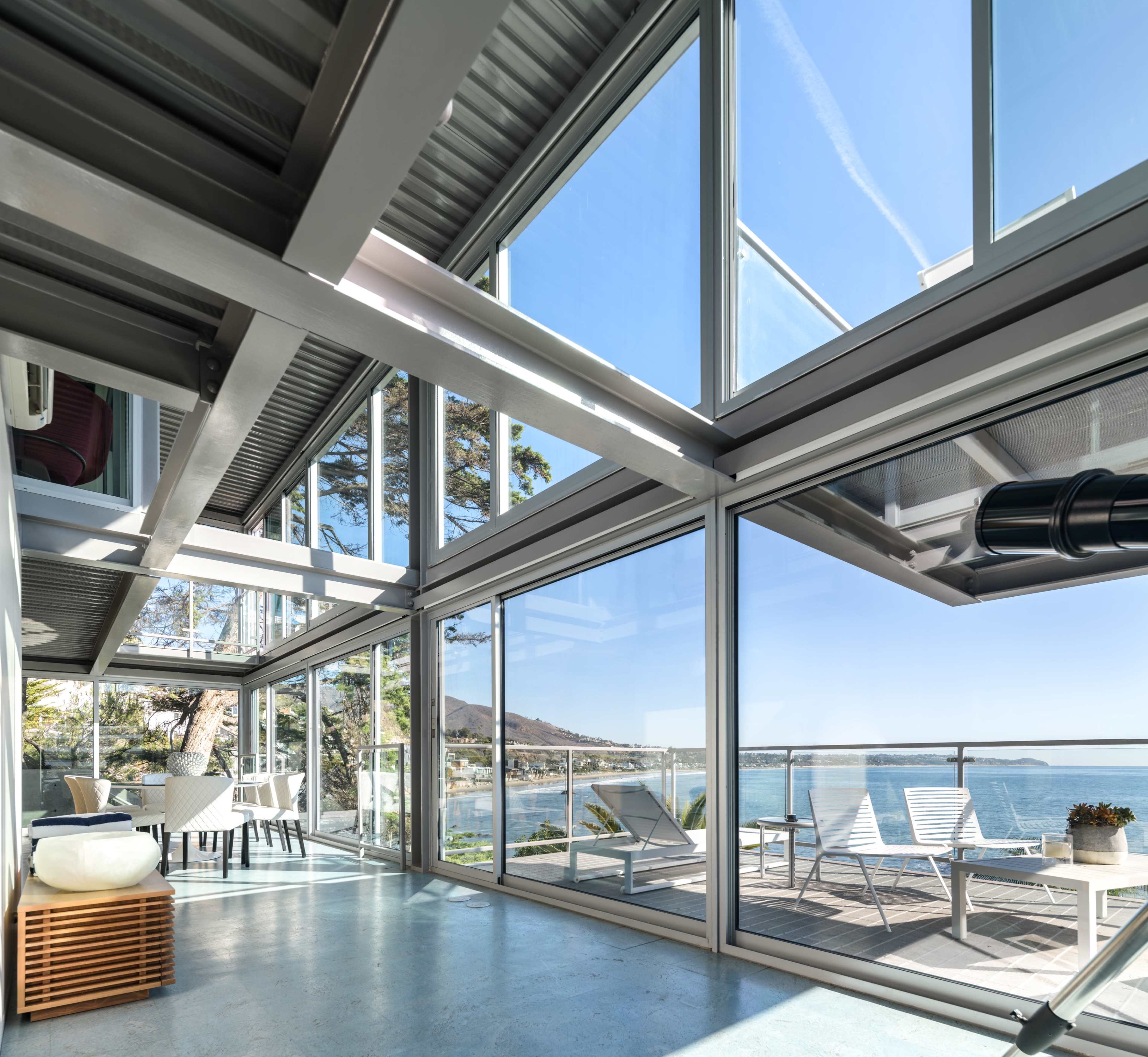 A modern glass-enclosed living space with a view of the ocean, featuring sleek furniture and a balcony with white lounge chairs.