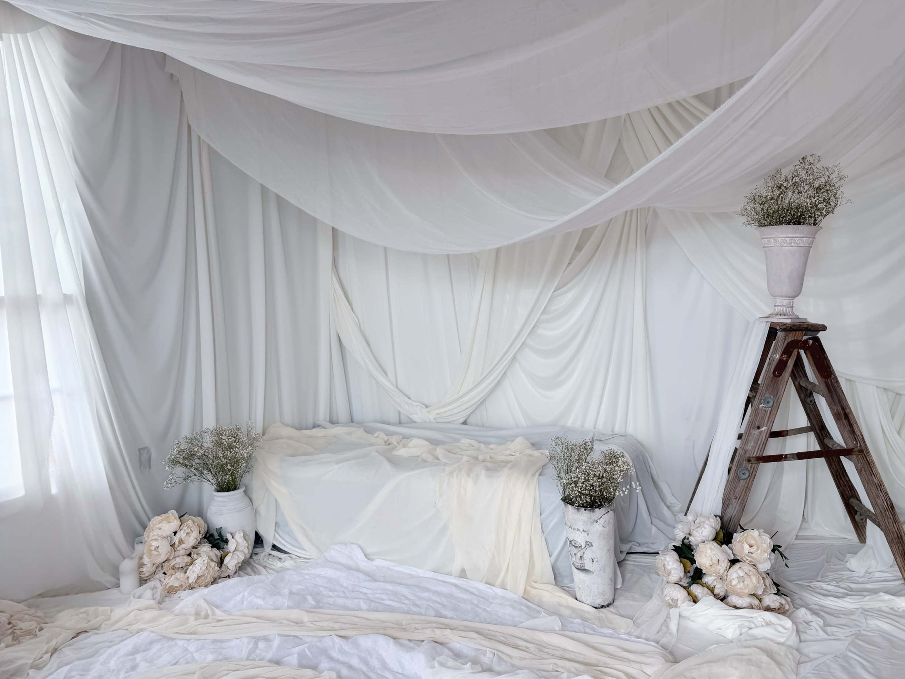 The image shows a room with a white draped backdrop, a light-colored sofa, and a wooden ladder adorned with a decorative vase of flowers.