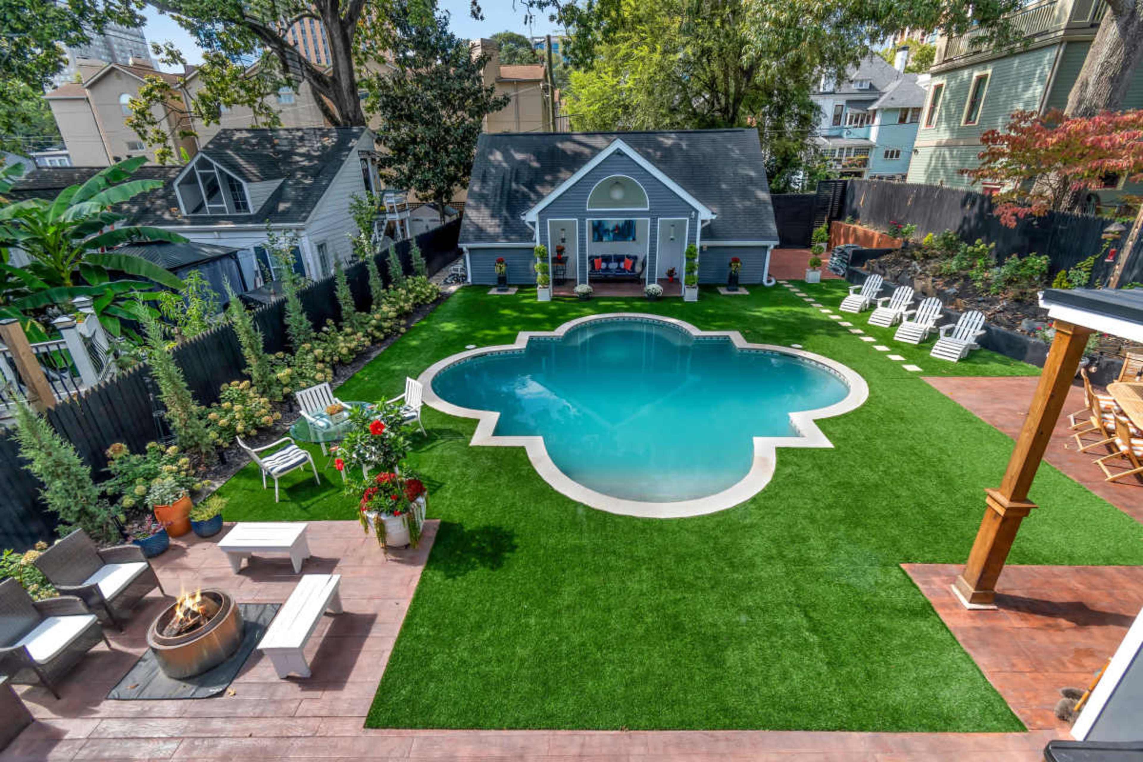 The image shows a backyard featuring a green synthetic lawn with a pool in the shape of a quatrefoil and seating areas surrounded by decorative plants.