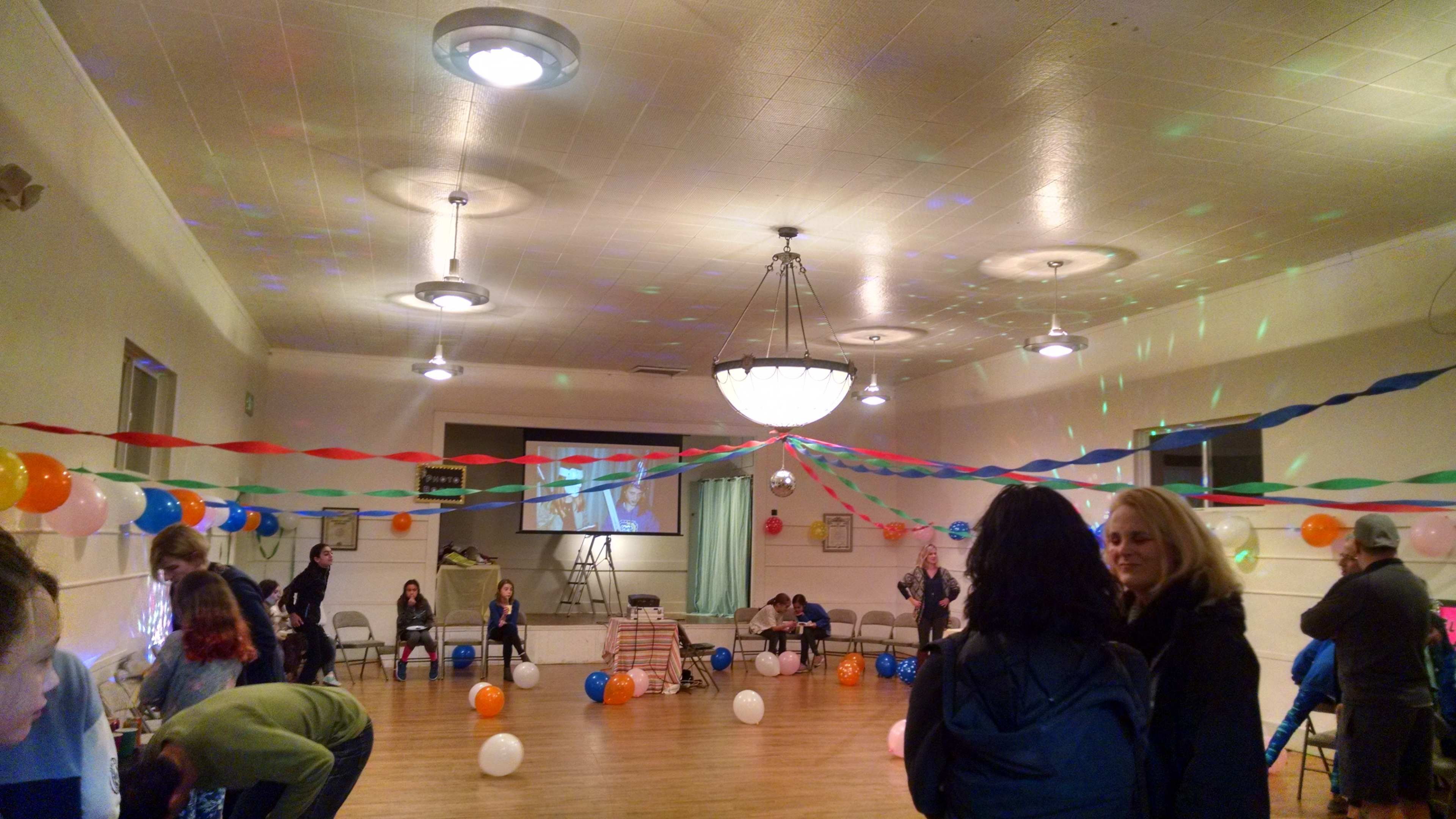 A decorated indoor event space features colorful balloons, streamers, and a few people mingling around.