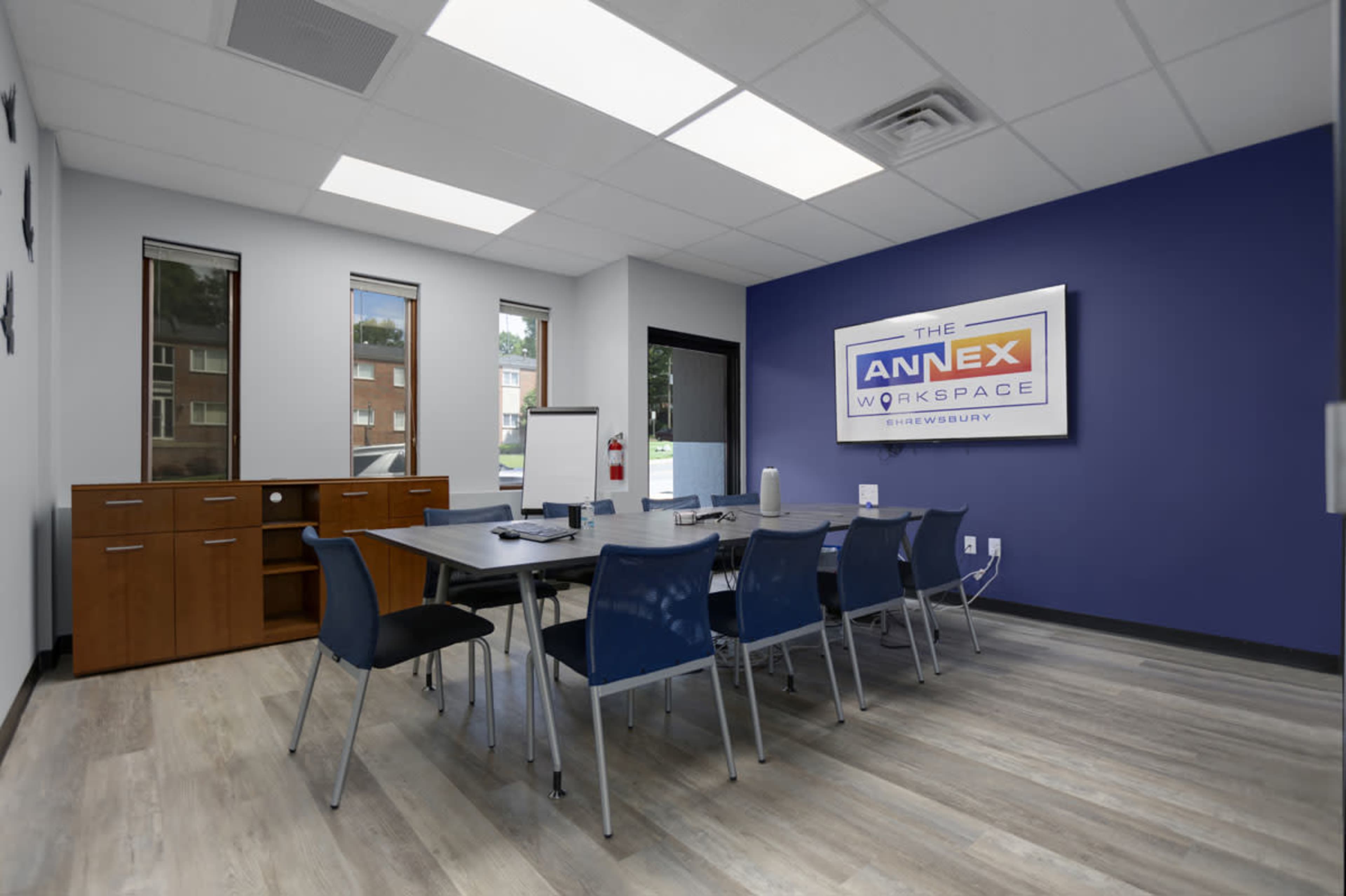 A modern conference room features a long table surrounded by blue chairs, with a large wall-mounted sign displaying "The Annex Workspace."