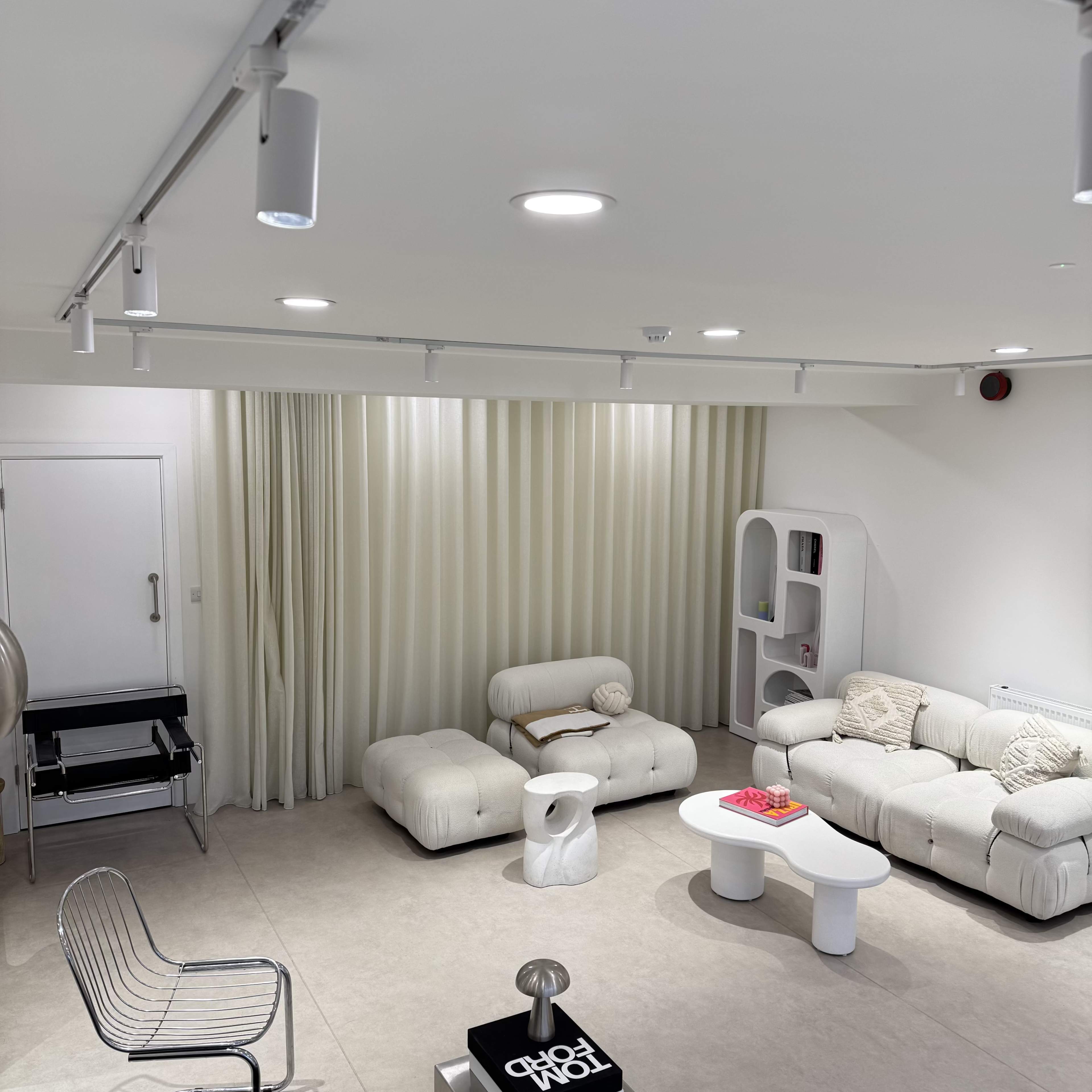 The image shows a modern living room with a white sofa, two cream-colored lounge chairs, a circular coffee table, and a decorative white pedestal paired with a glass shelf against a backdrop of light curtains.