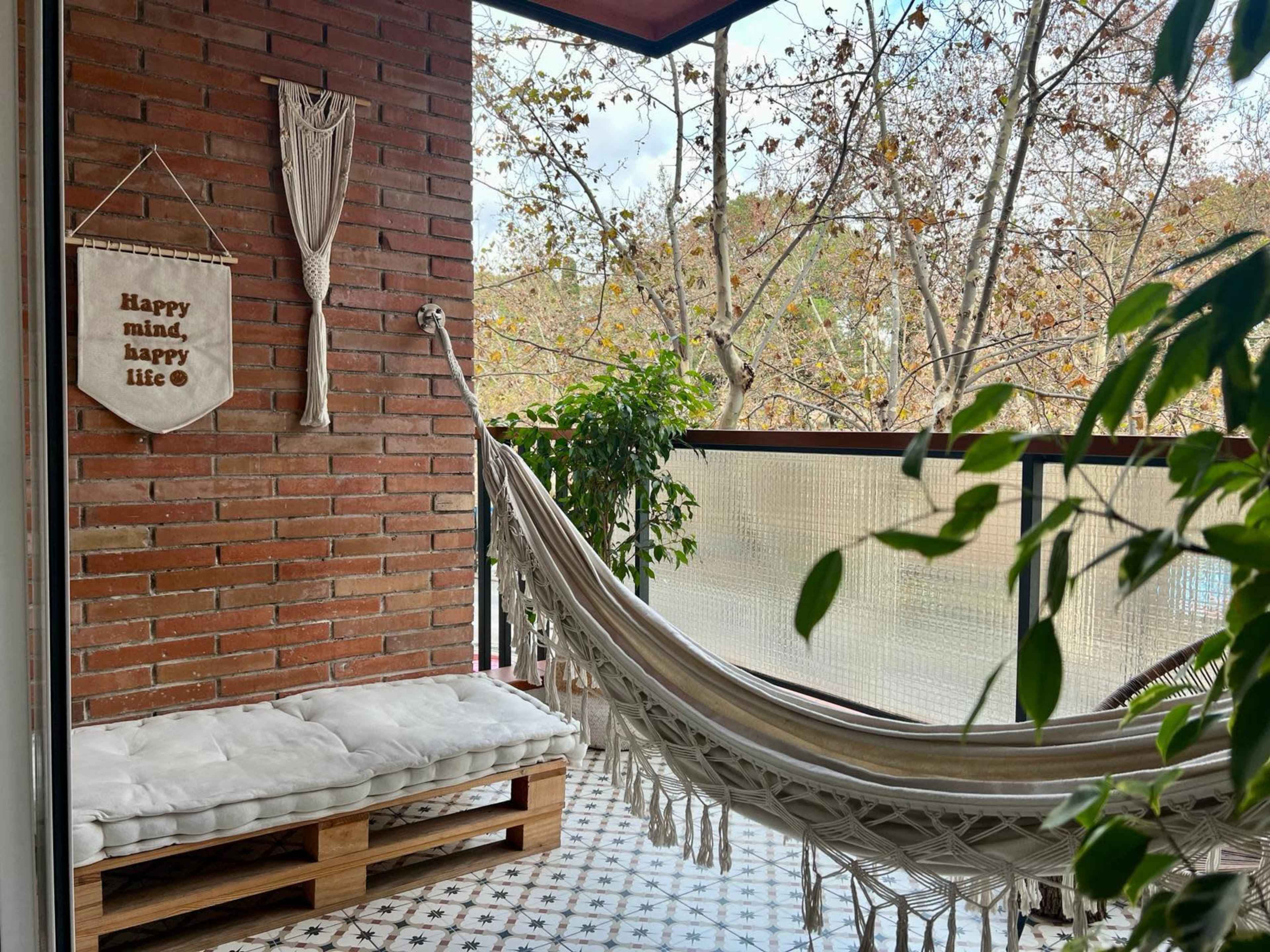 A cozy balcony features a hammock, a bench made of pallets, and plants, with a decorative wall hanging that reads "Happy mind, happy life."