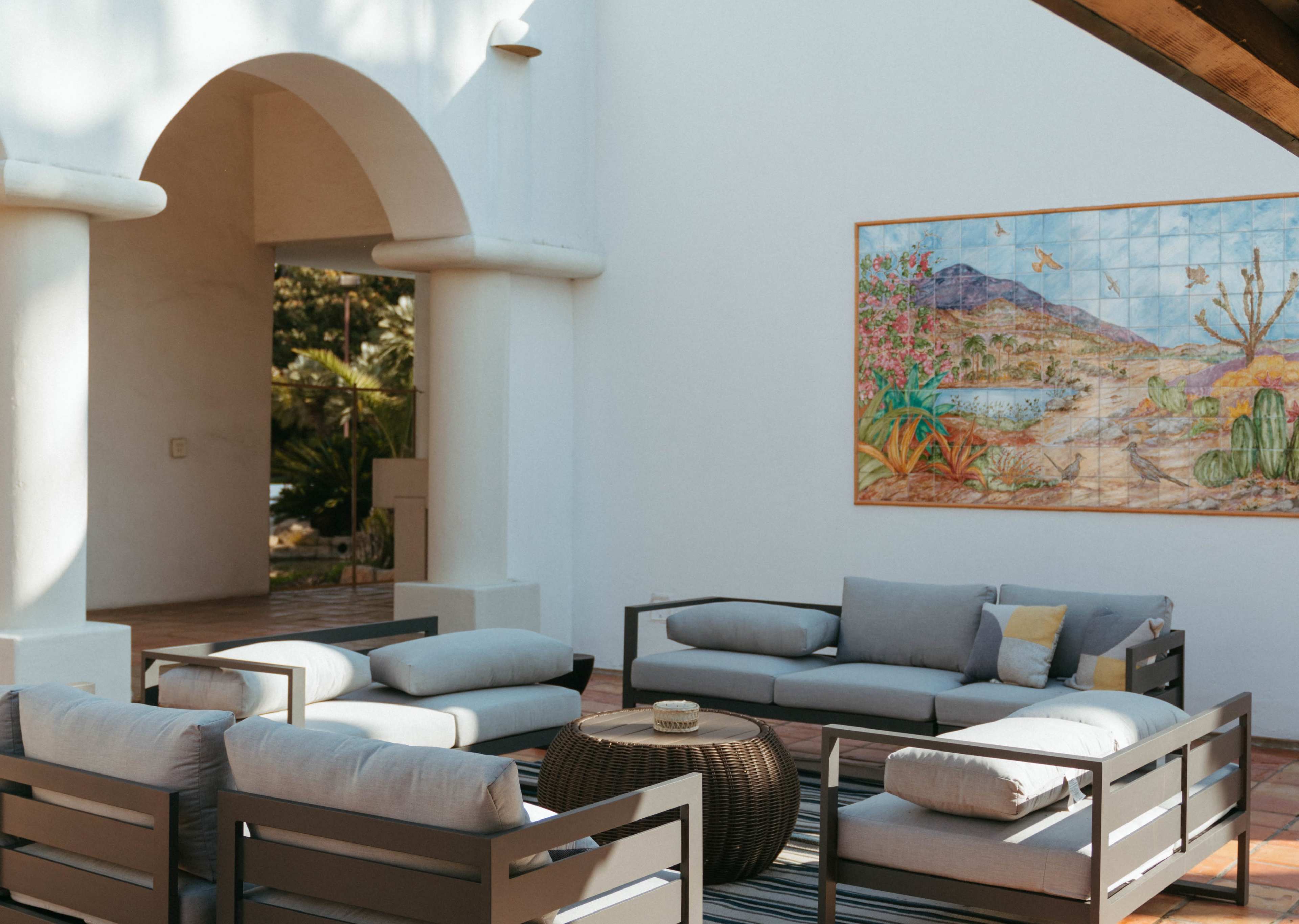 A spacious outdoor seating area features gray sofas arranged around a circular table, with a large mural of a desert landscape on the wall.