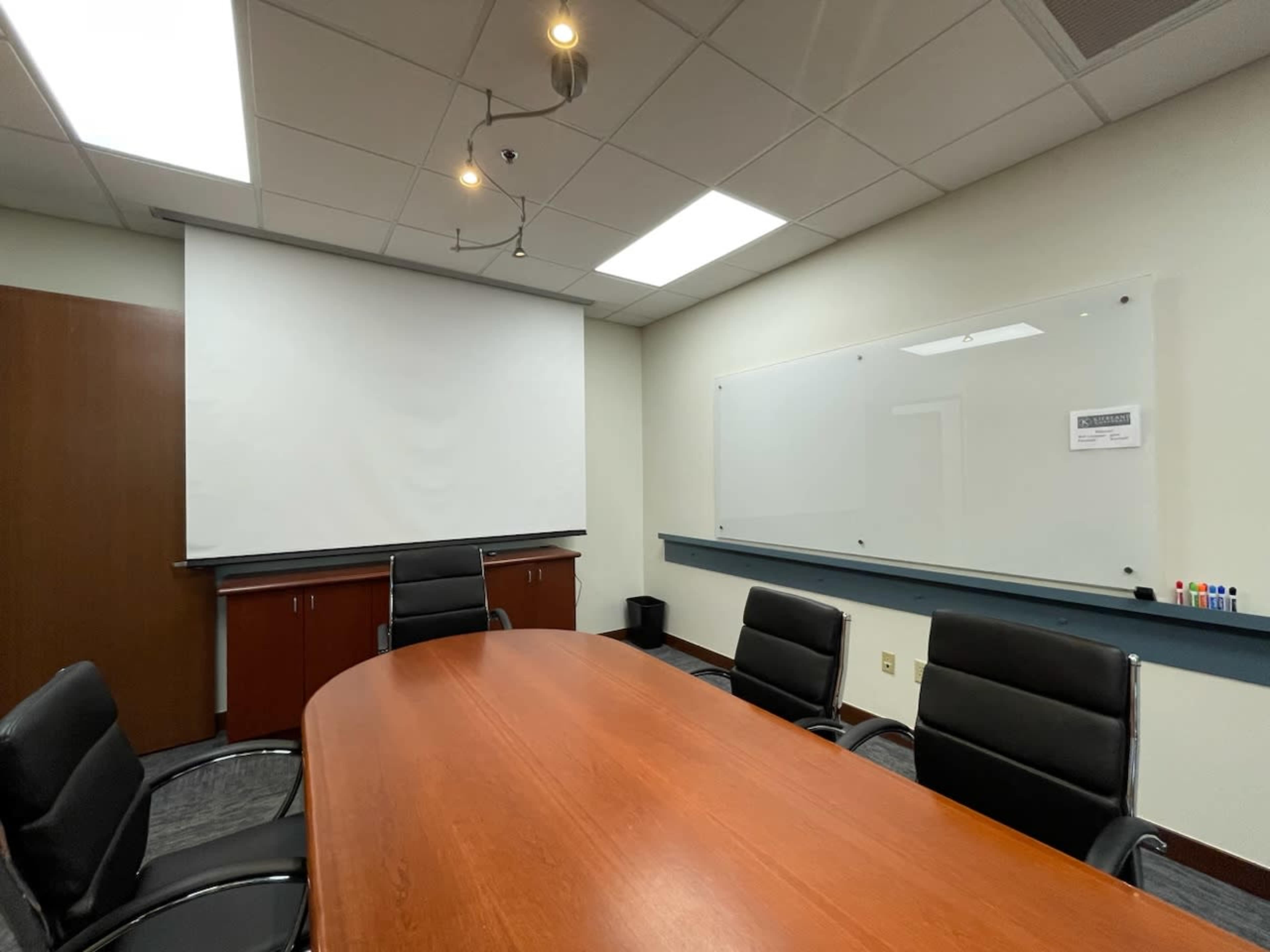 A conference room with a wooden oval table surrounded by black chairs, a projection screen, and a whiteboard on the wall.