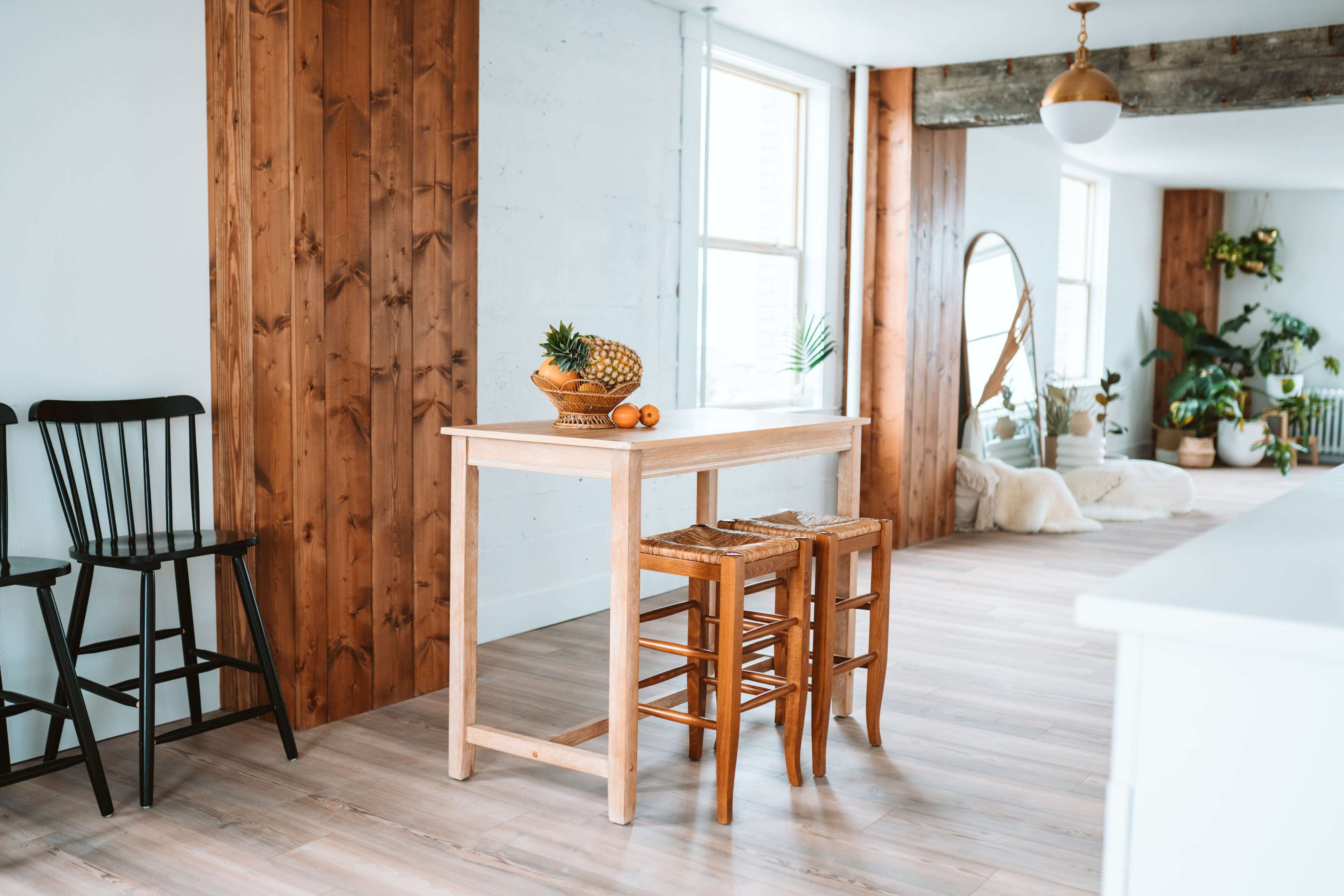 A wooden table with two stools is placed in a brightly lit room featuring a mix of natural wood and white walls, with plants and decor visible in the background.