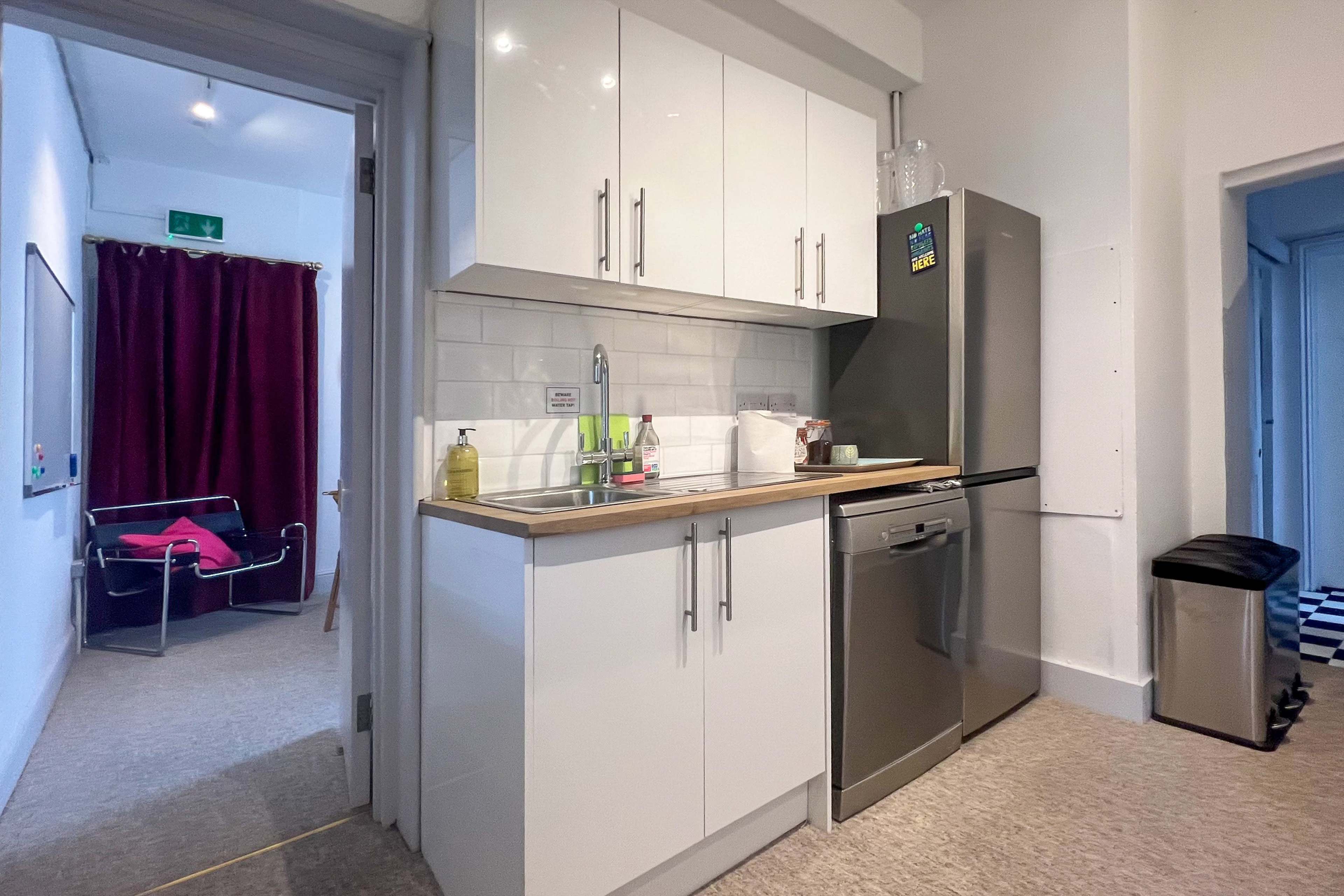 A compact kitchen area featuring white cabinets, a stainless steel refrigerator, a sink, and a countertop, with a doorway leading to a room with burgundy curtains in the background.