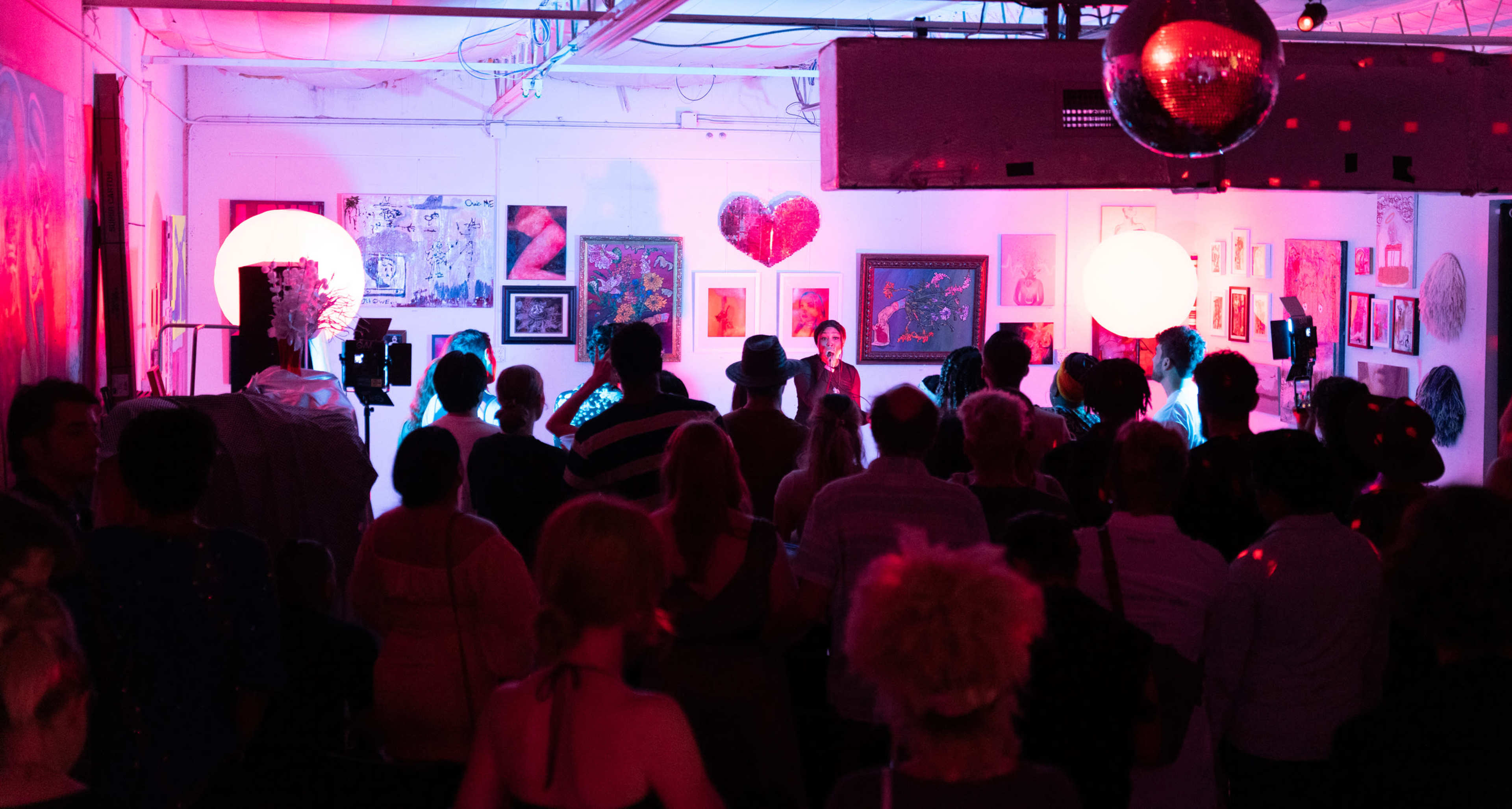 A crowd gathers in an art gallery, with illuminated artworks displayed on the walls and large glowing orbs adding to the ambiance.